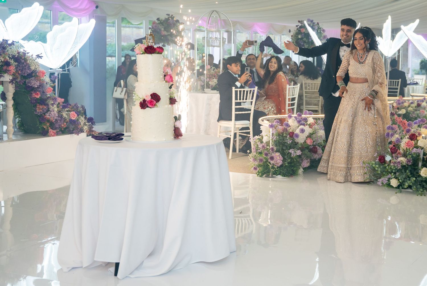 Couple cutting a white tiered wedding cake with sparklers at an elegant floral reception in a marquee.