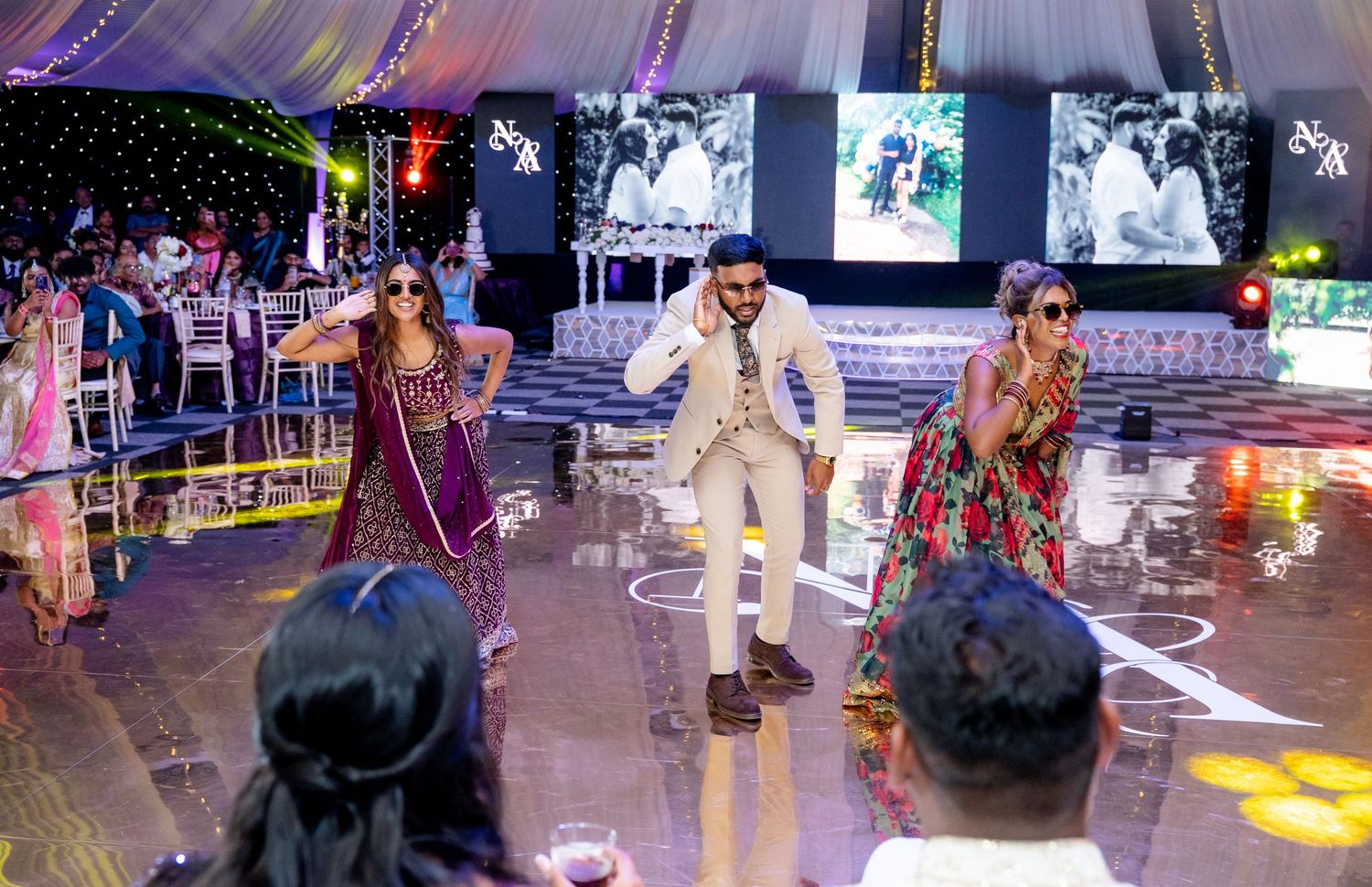 Three performers dancing at an elegant Indian wedding reception with decorative lighting and guests seated in background.