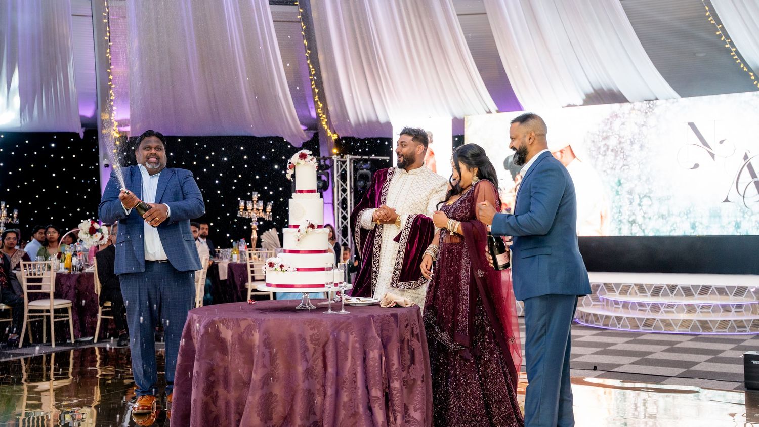 Indian bride and groom cutting wedding cake with family members gathered around in decorated reception hall.