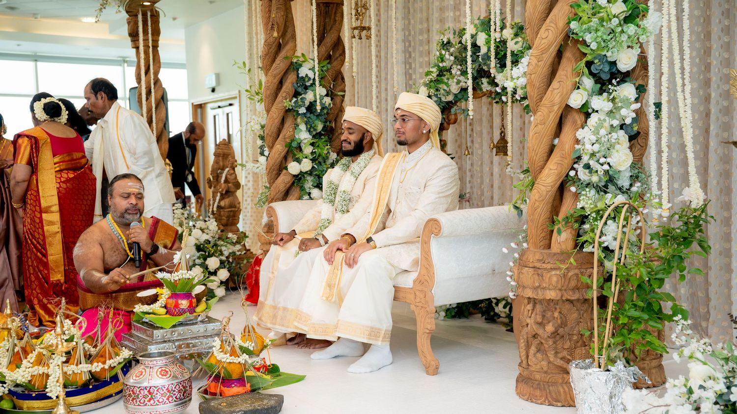 Two grooms in white sit at a beautifully decorated Hindu wedding mandap with floral arrangements.