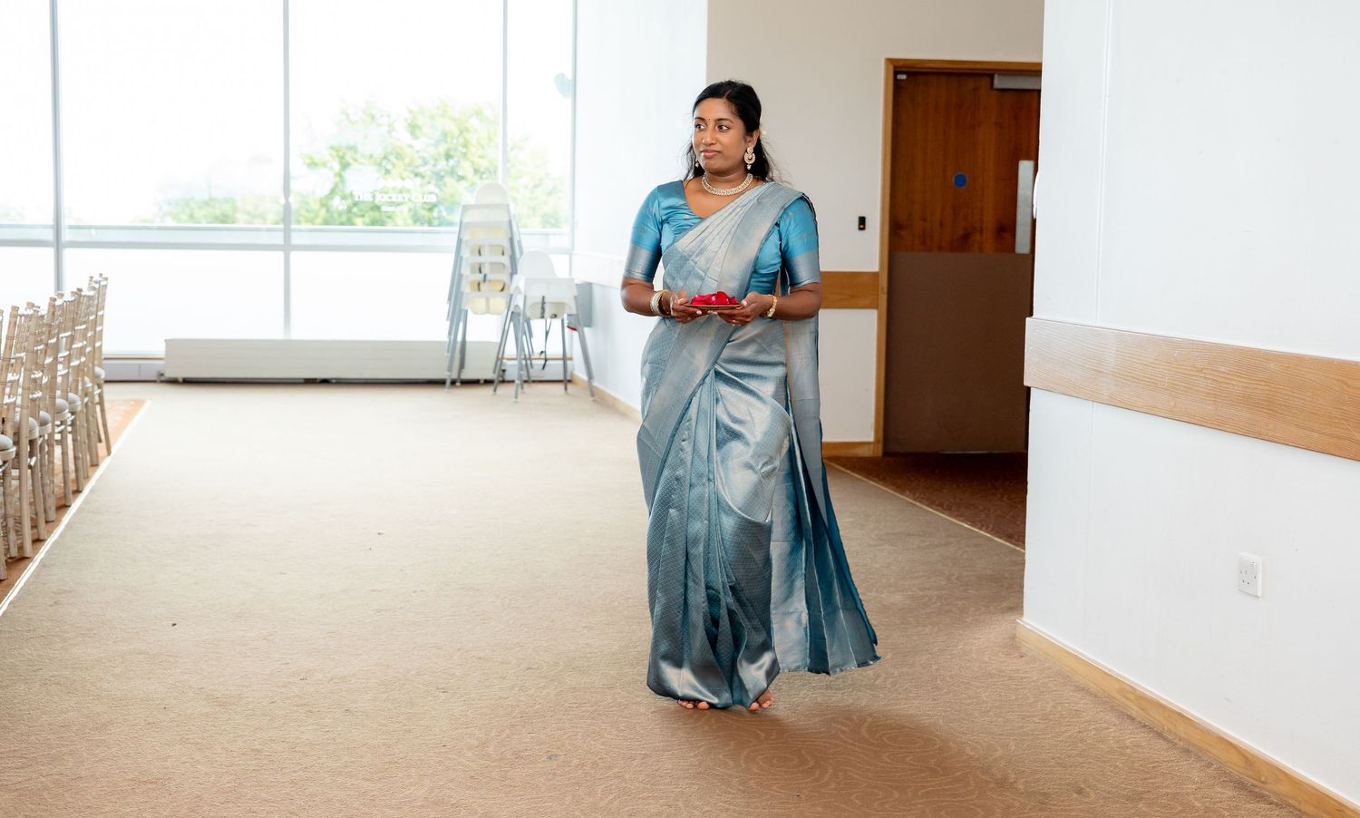 Woman in blue-grey silk saree walking through bright wedding venue hall carrying ceremonial red plate.