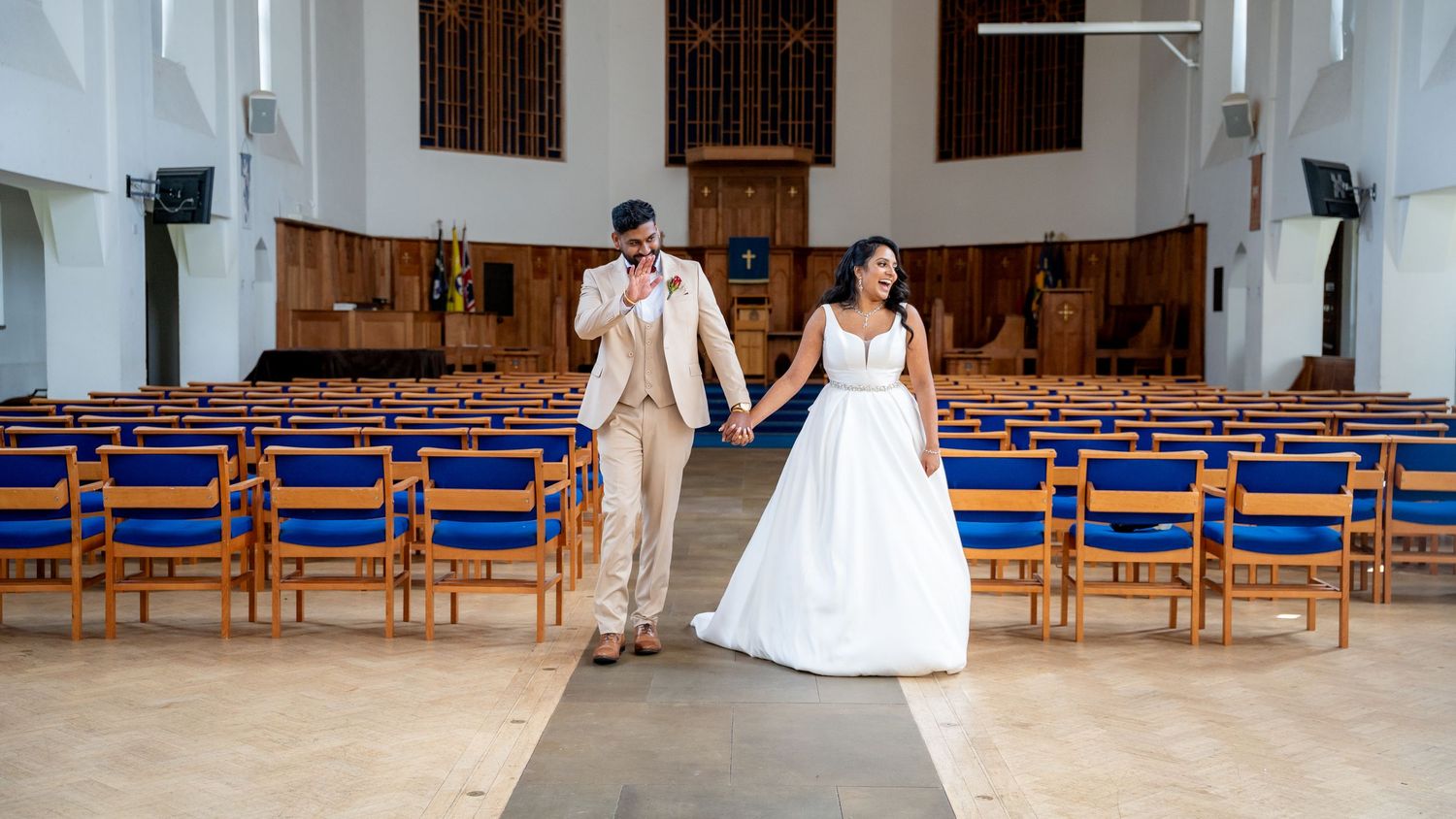 Bride and groom walk hand-in-hand down the center aisle of a modern church with blue-cushioned pews.