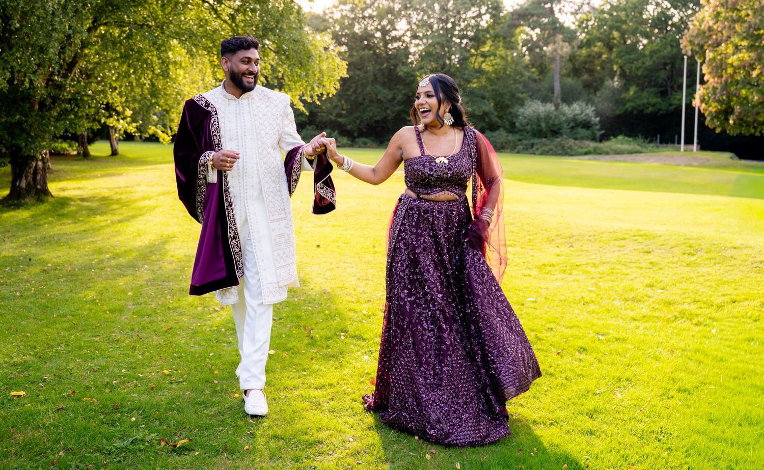 Indian couple in traditional attire walking on green lawn, man in white sherwani, woman in purple lehenga.