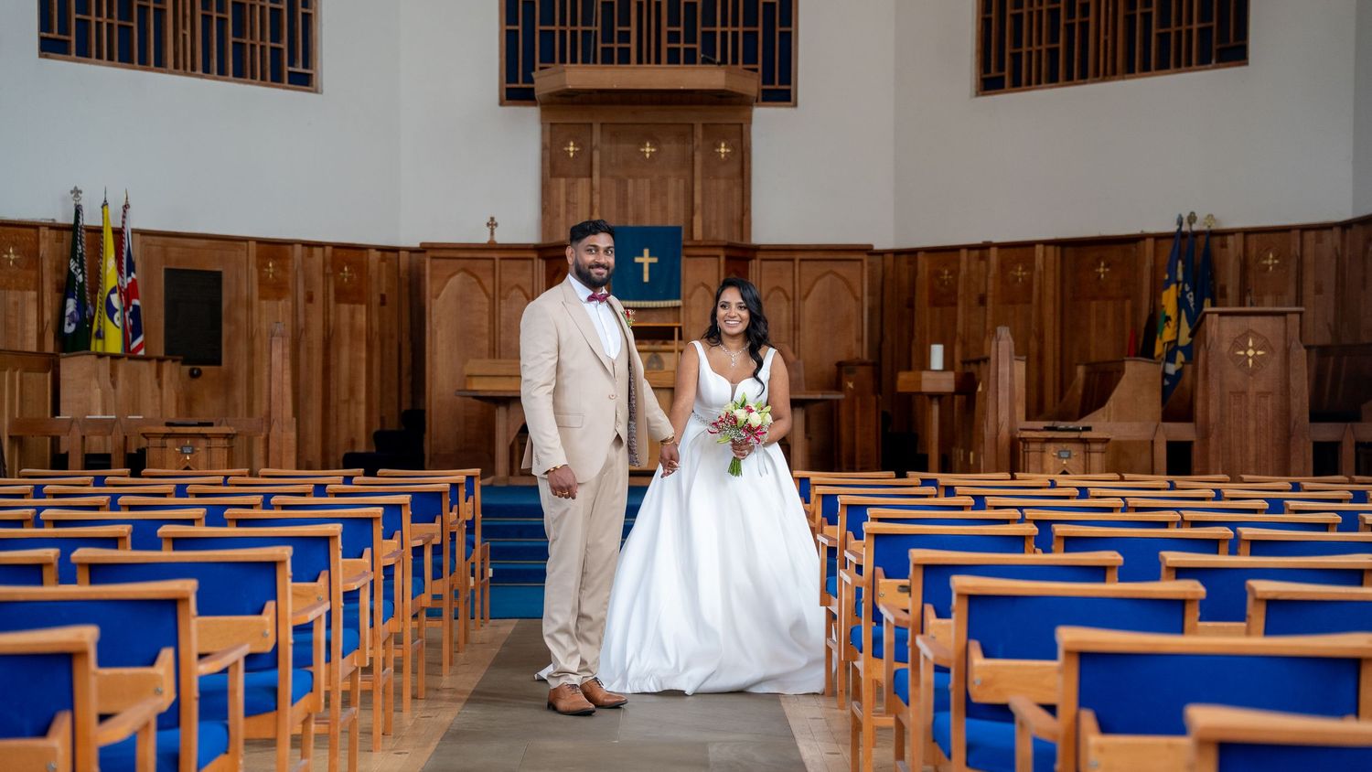 Bride and groom pose together at church altar, she holds a bouquet, surrounded by blue pew seating.