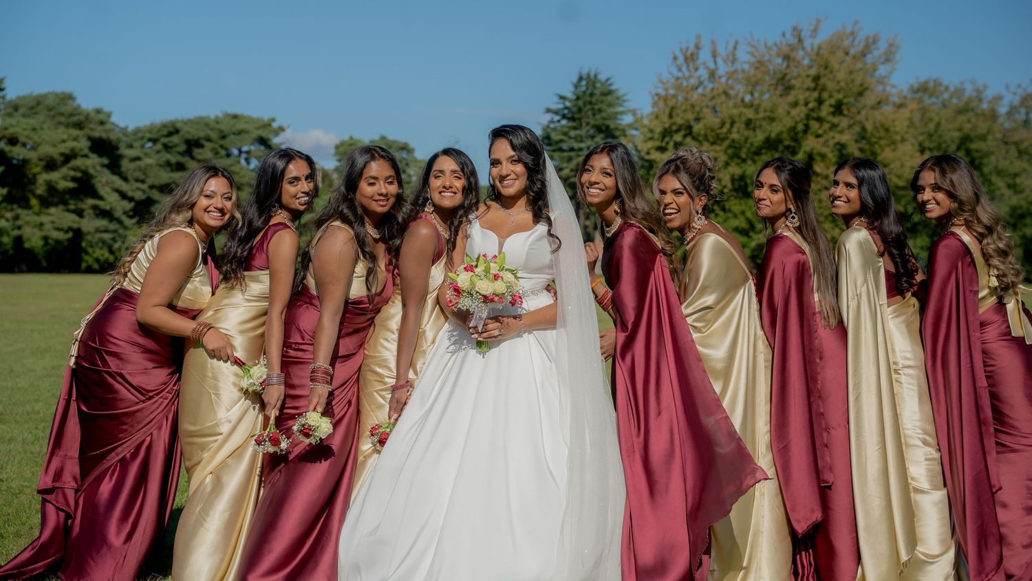 Bride in white gown poses with bridesmaids in burgundy and gold satin dresses outdoors on a sunny day.