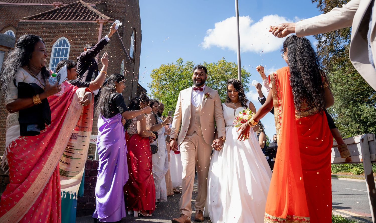 Indian wedding couple walks through cheering guests in colorful saris outside a brick church on a sunny day.