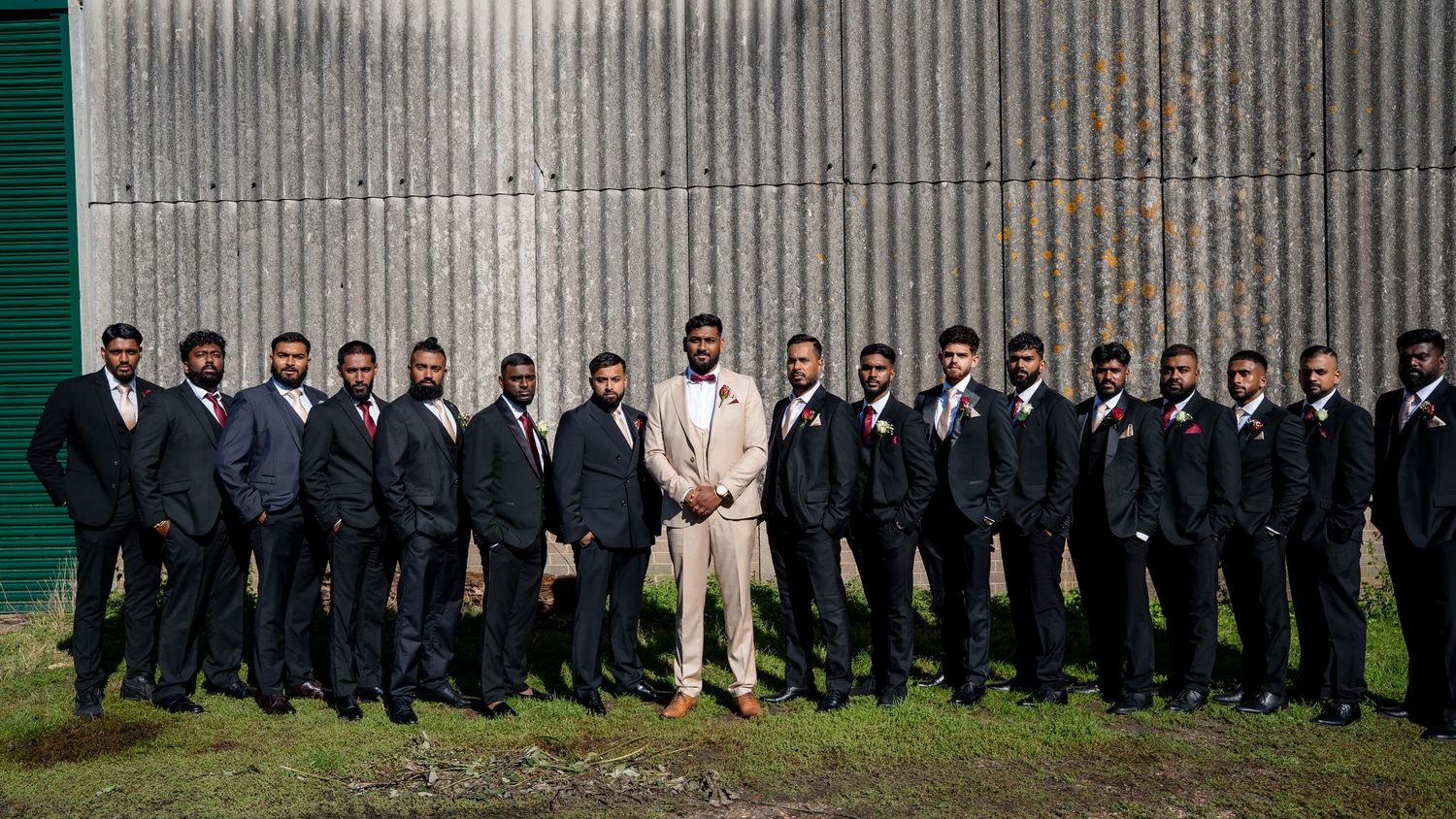 Large wedding party with groom in beige suit and groomsmen in navy suits pose outdoors.
