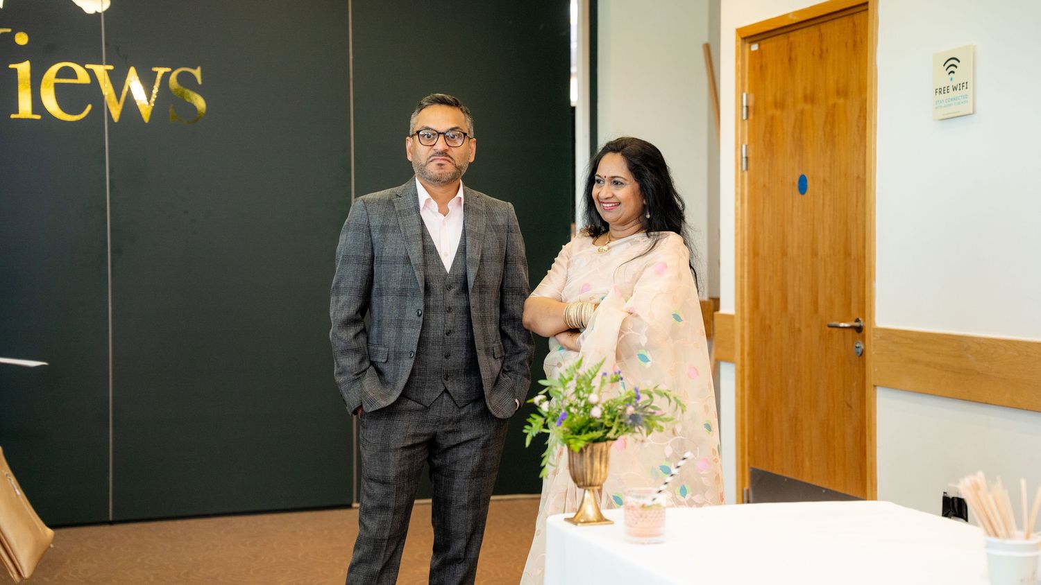 Couple in formal attire posing together at an indoor wedding venue near a floral arrangement.