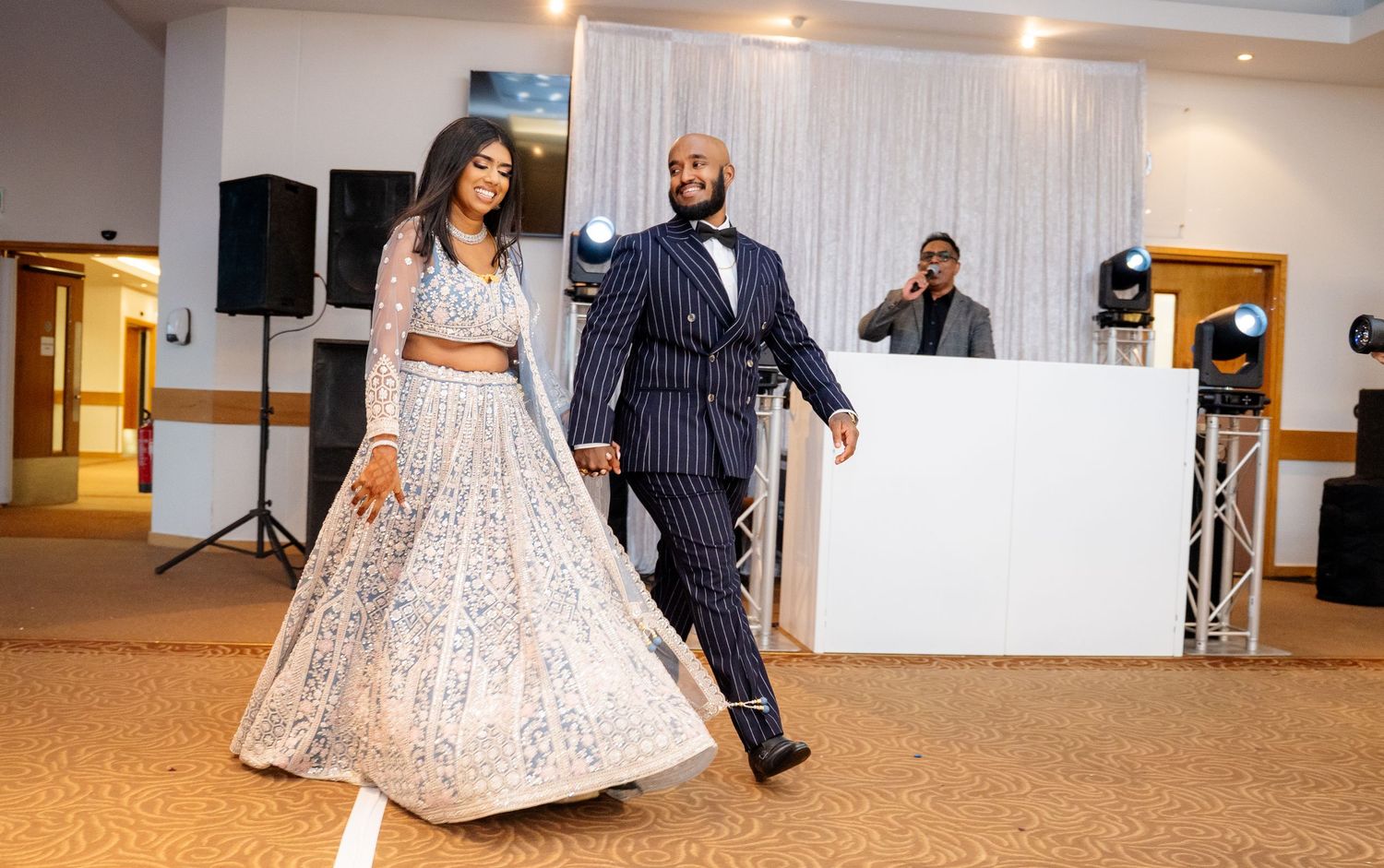 Bride in silver lehenga and groom in navy pinstripe suit dance together at wedding reception.