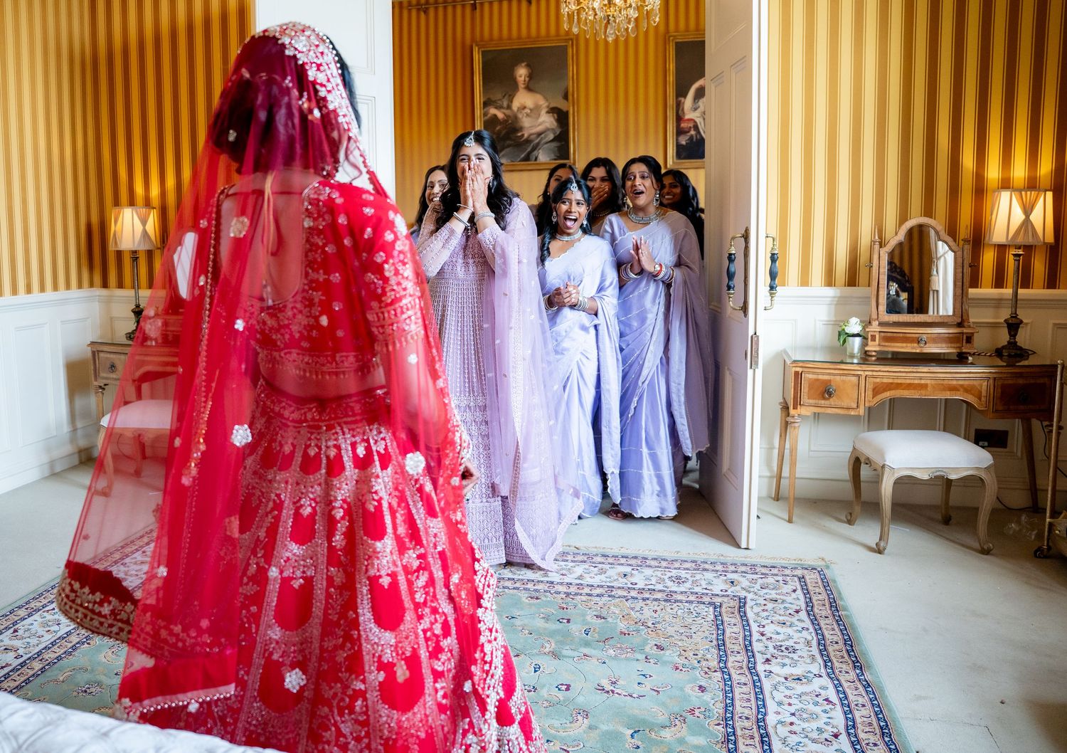 Indian bride in red lehenga revealed to amazed bridesmaids in lavender dresses in elegant room.