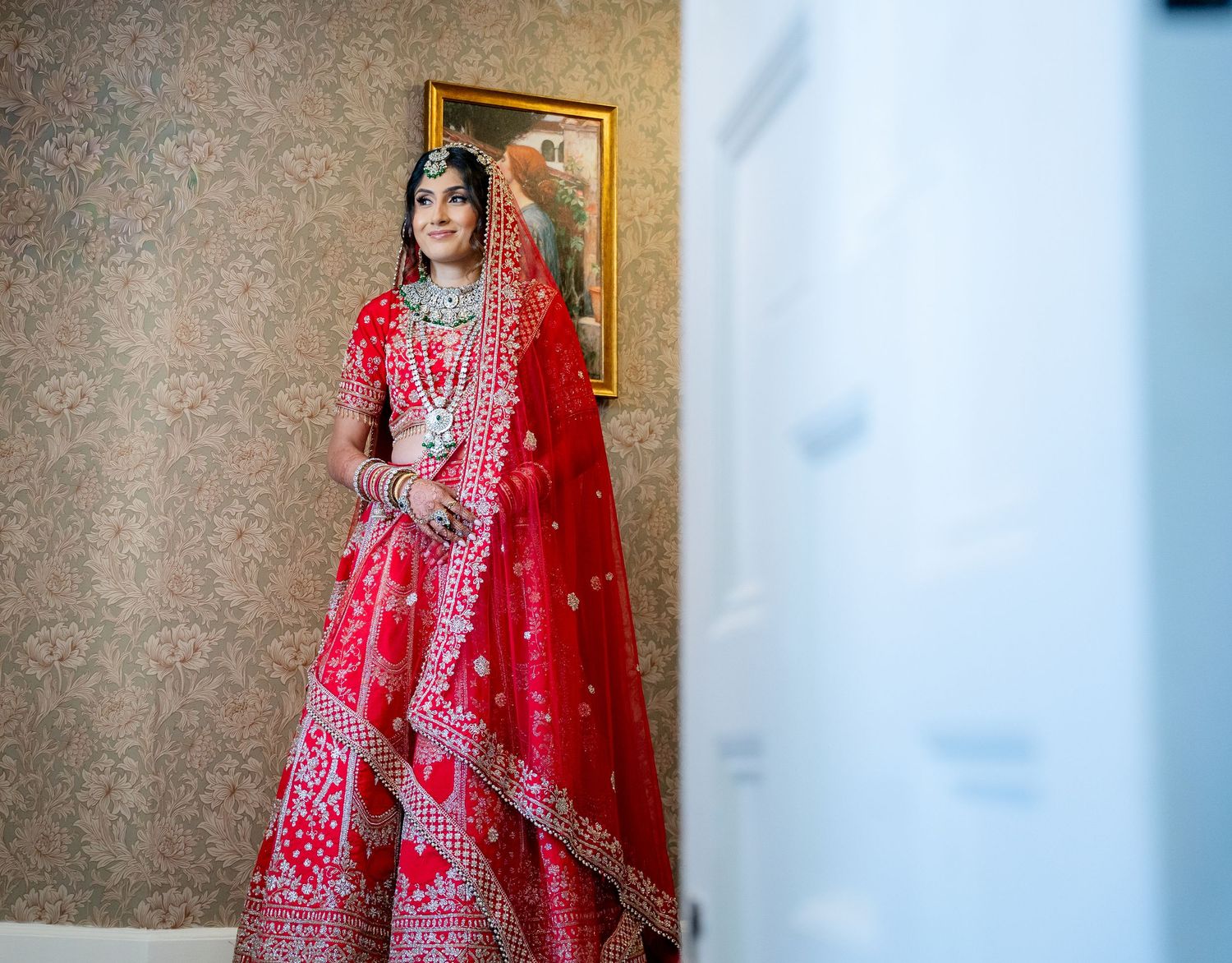 Indian bride in red embroidered lehenga standing gracefully in elegant wallpapered dressing room.