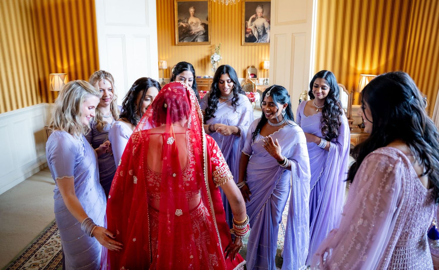 Indian bride in red lehenga surrounded by bridesmaids in lavender attire.