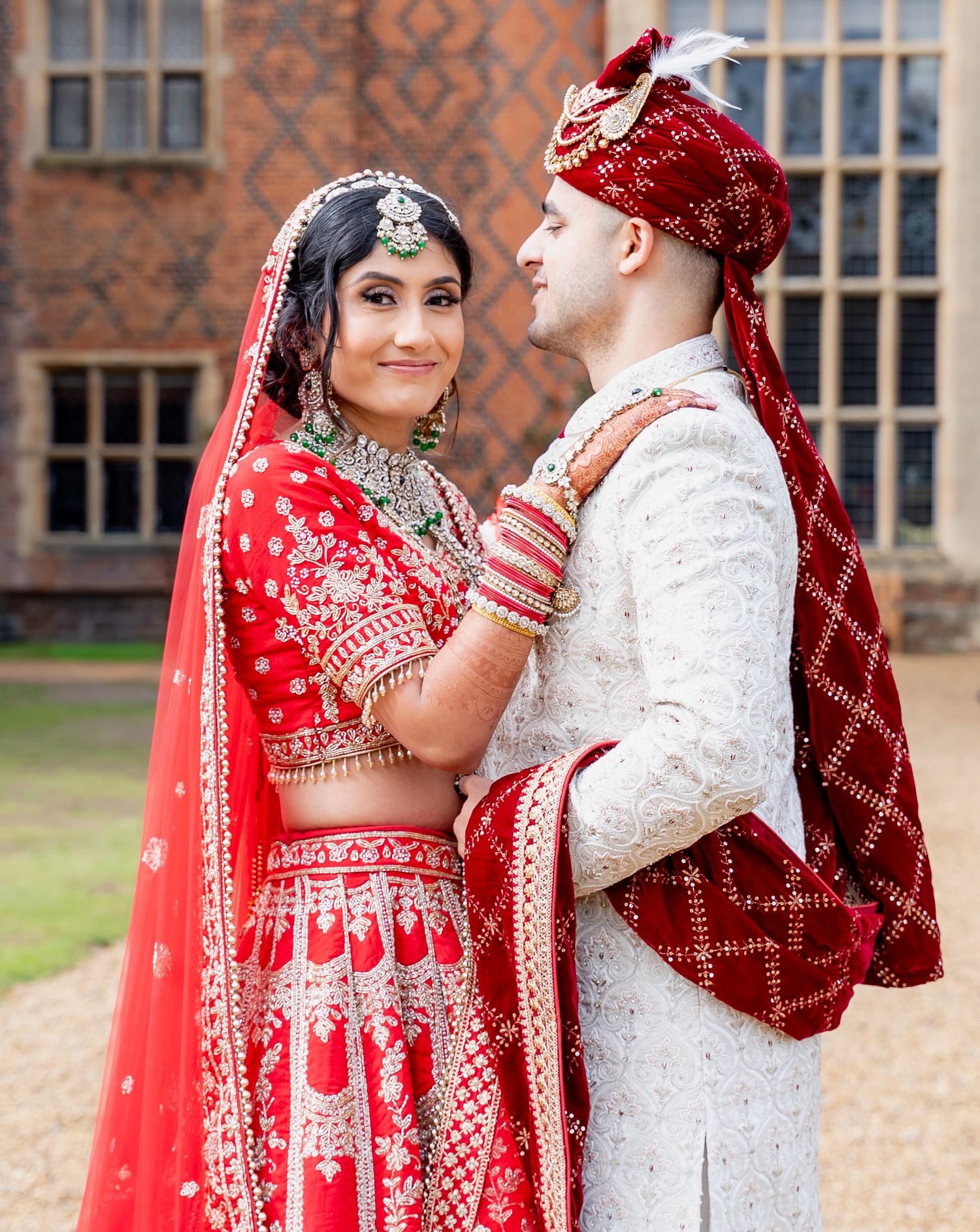 Indian bride in red bridal lehenga and groom in white sherwani posing together outside grand estate.