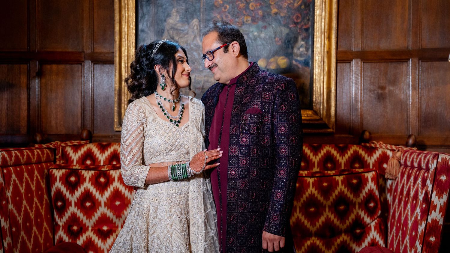 South Asian couple in traditional wedding attire share a romantic moment in wood-paneled room.