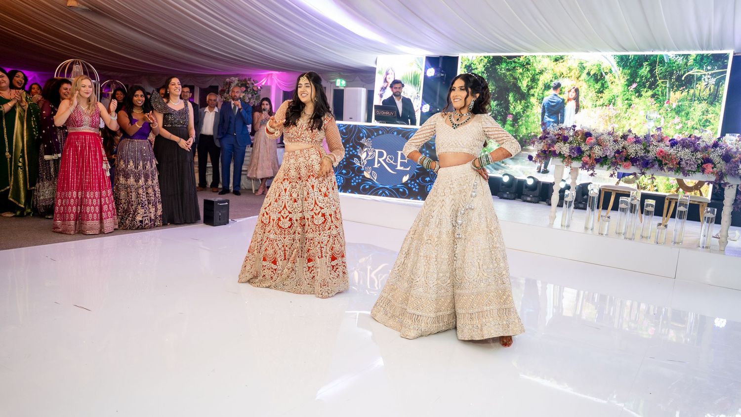 Two women in embroidered lehengas perform an elegant dance on a white dance floor at a wedding.