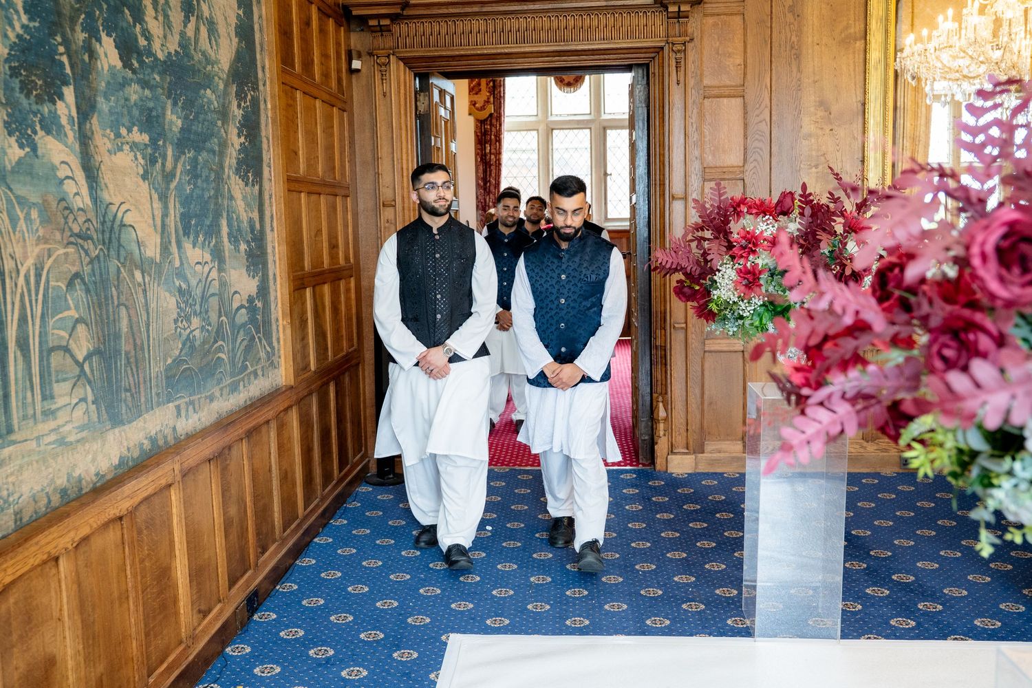 Men in traditional shalwar kameez walking through ornate wooden doorway with floral decor.