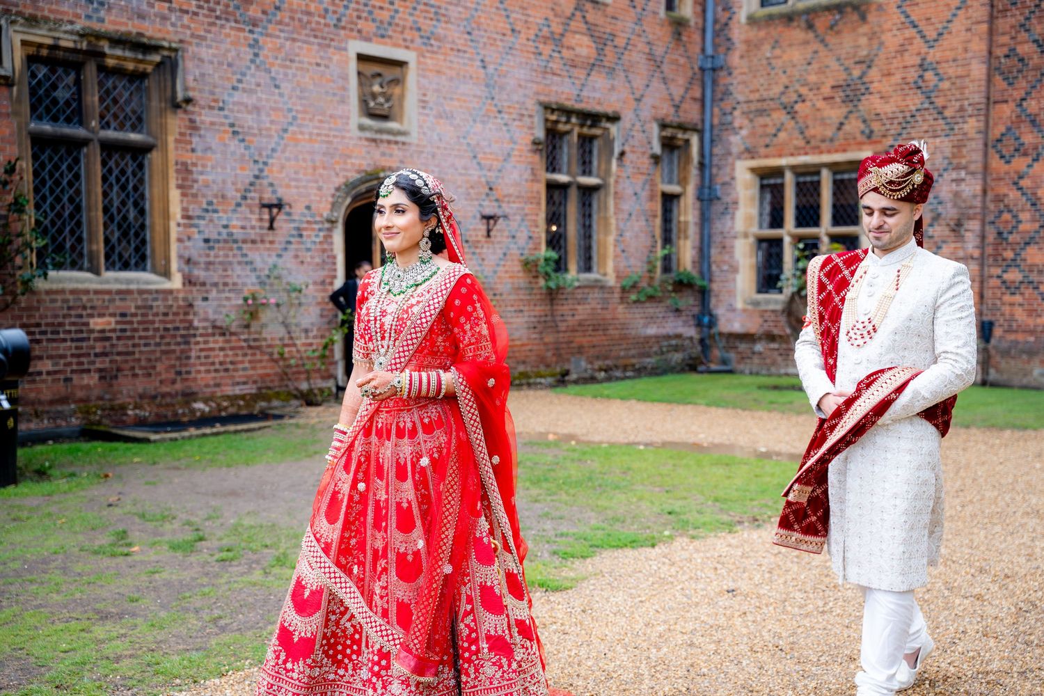 Indian bride in red lehenga and groom in white sherwani walking outdoors at a historic venue.