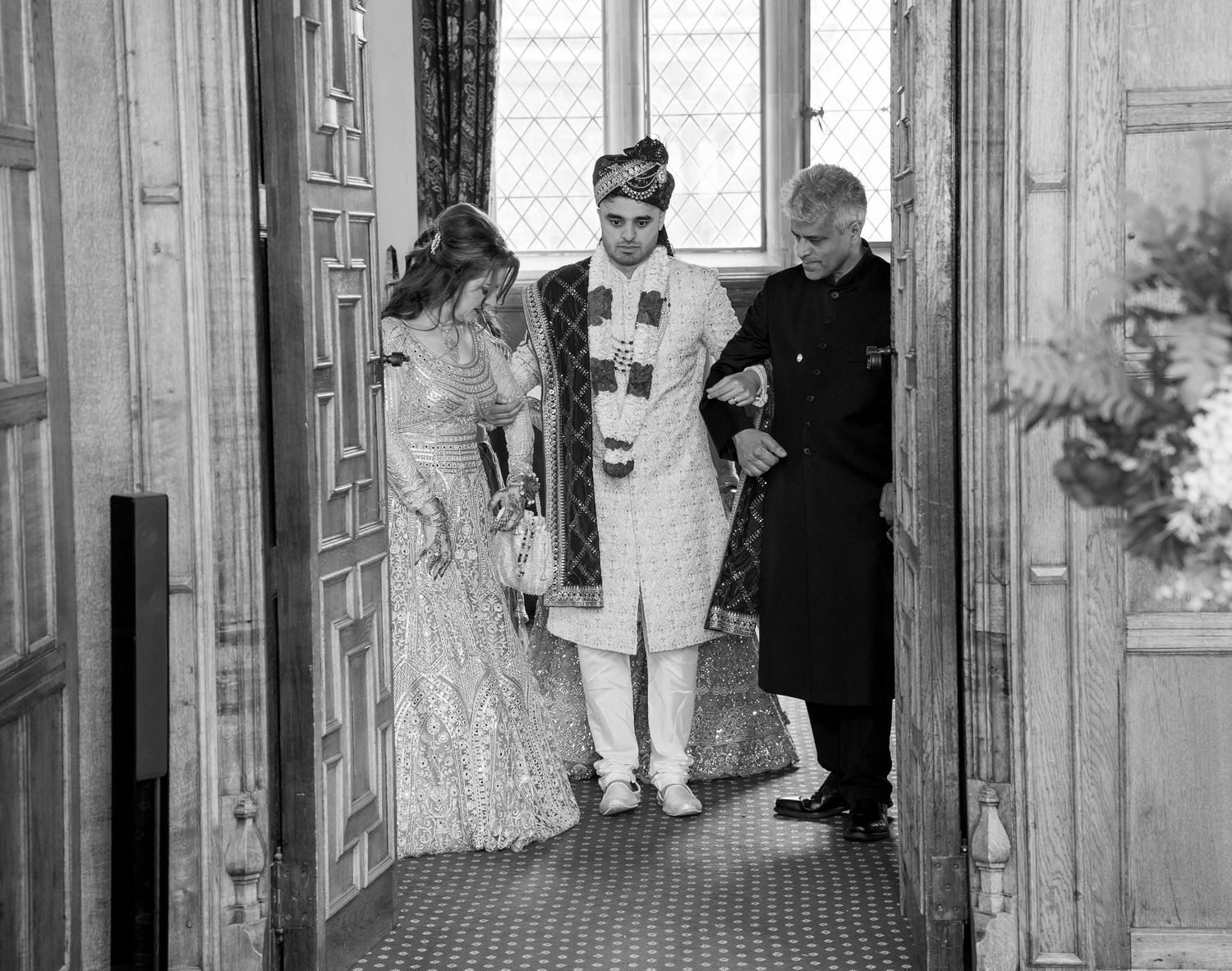 Groom in white sherwani enters with family during Indian wedding ceremony in elegant wood-paneled hall.