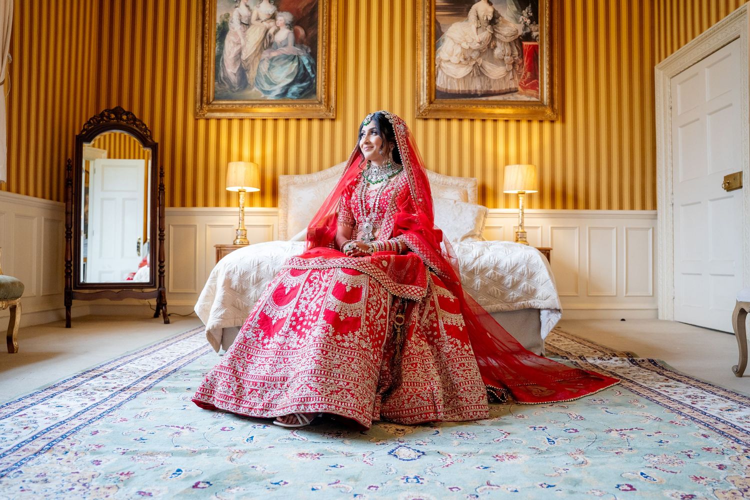 Beautiful Indian bride in red bridal lehenga sits on bed in ornate yellow room.