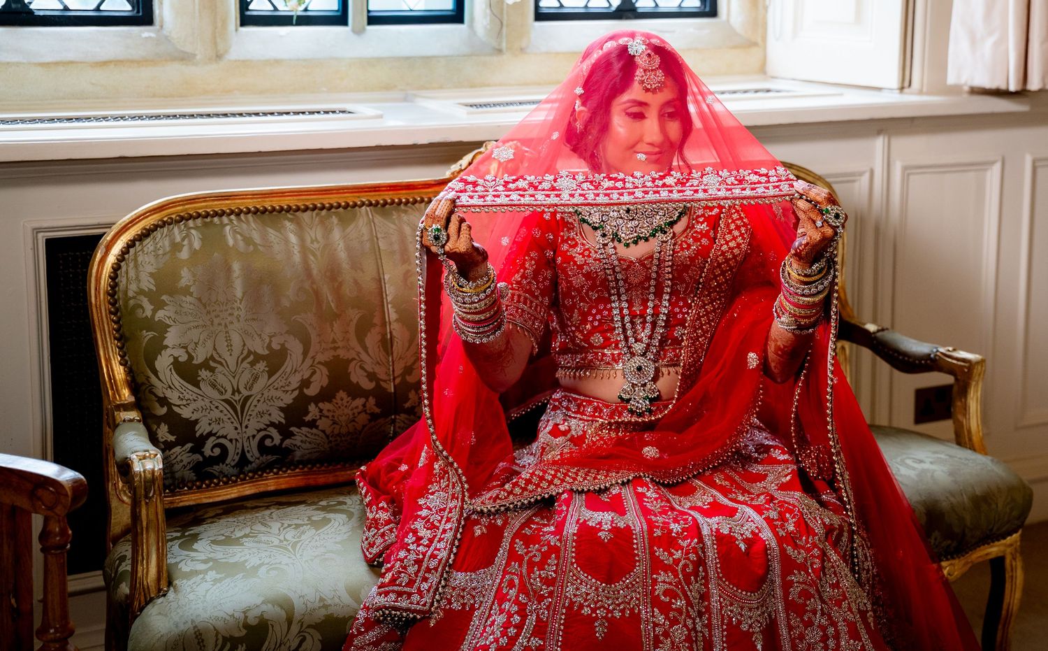 Indian bride in red bridal lehenga sits elegantly on antique gold sofa near window.