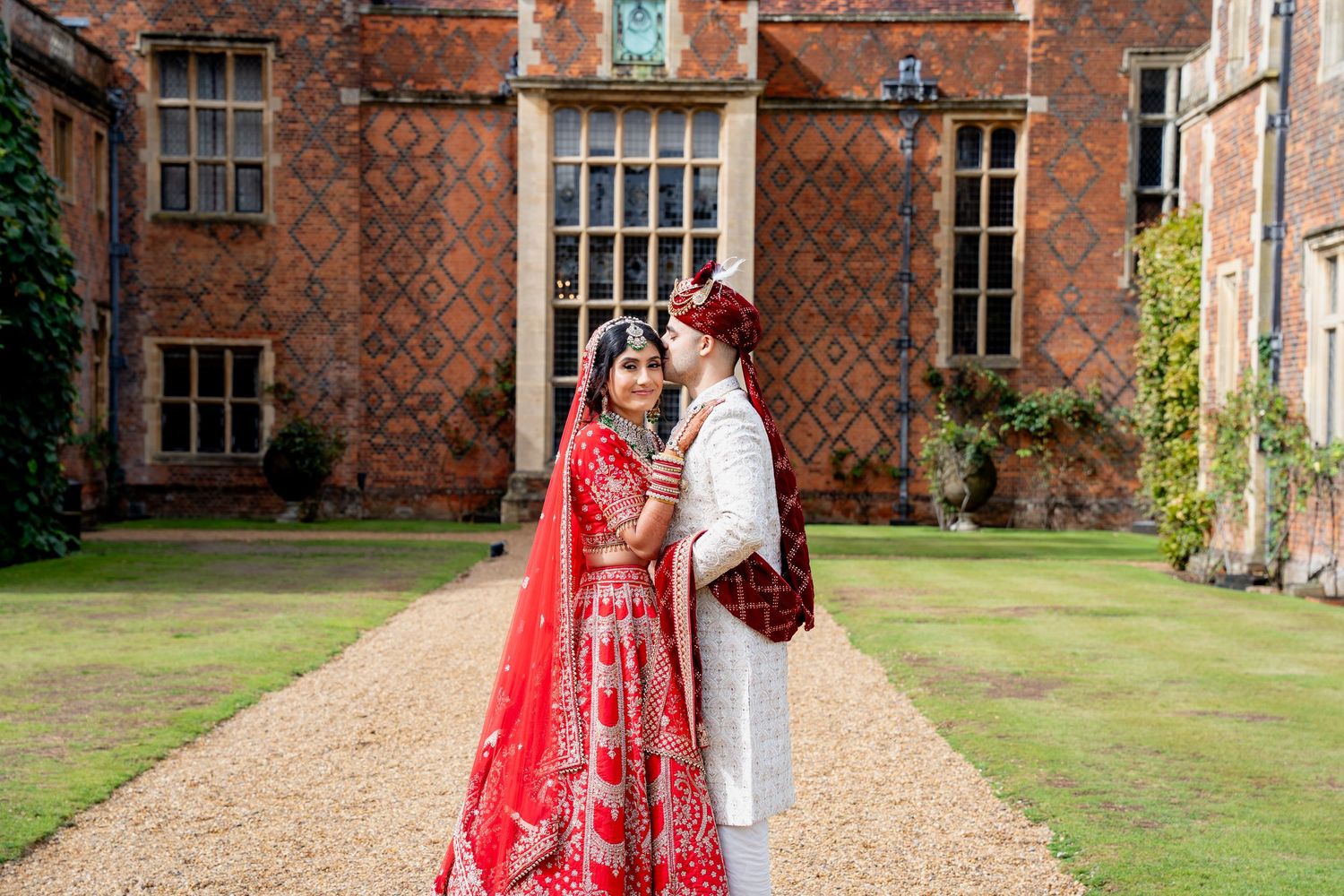 Indian wedding couple in vibrant red lehenga and white sherwani standing before a grand historic brick manor house.