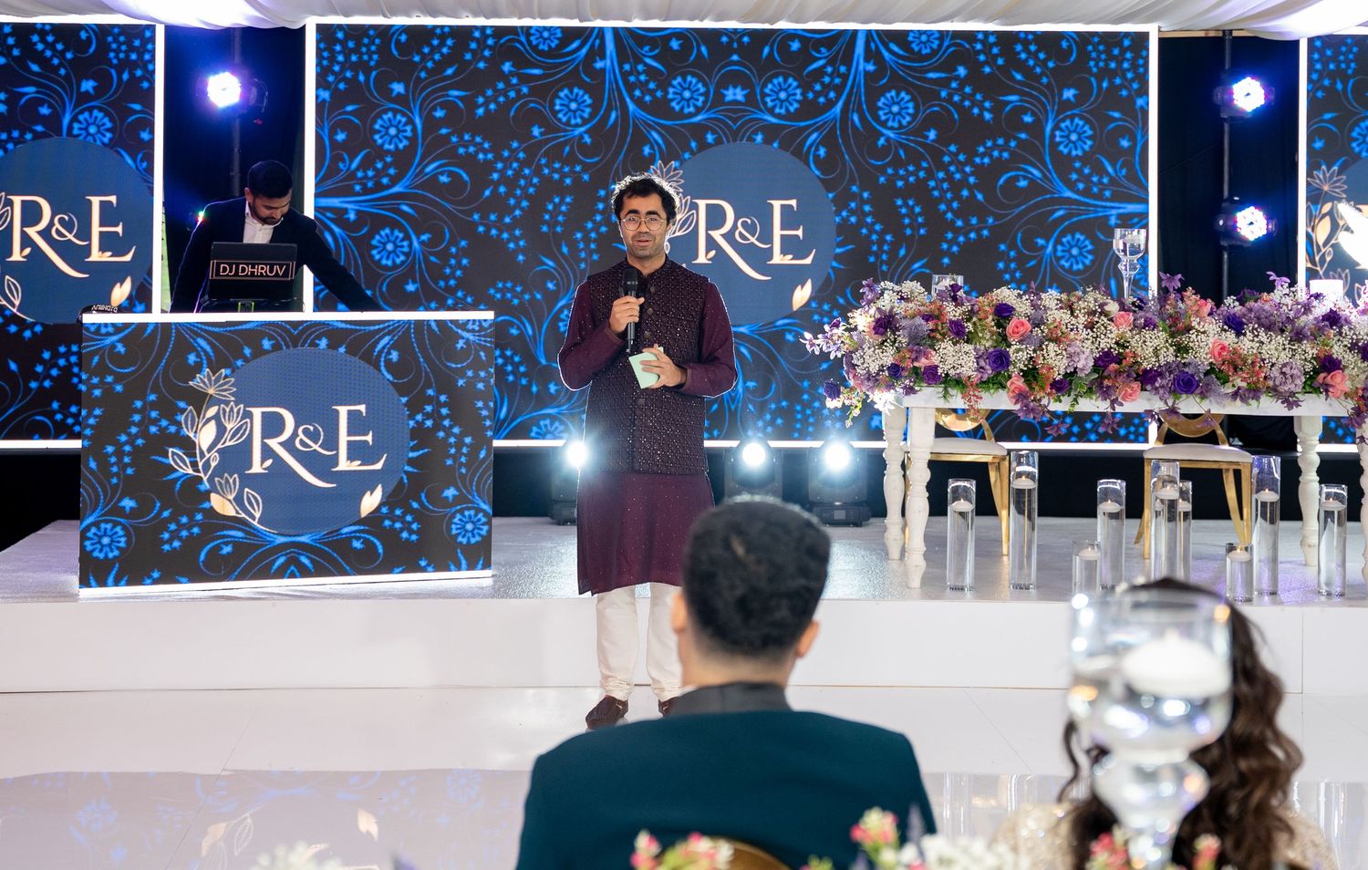 A speaker presents at an elegant event with blue floral backdrop and RE branding while attendees watch from seated tables.