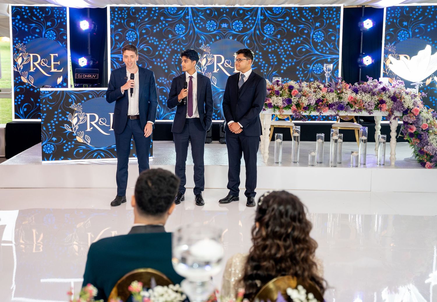 Three men in suits giving speeches at elegant wedding reception with floral blue R&E monogram backdrop.