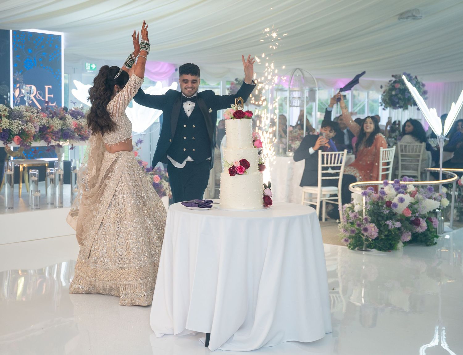 Bride and groom celebrate at wedding reception, raising hands near elegant white tiered cake with sparklers in decorated tent venue.