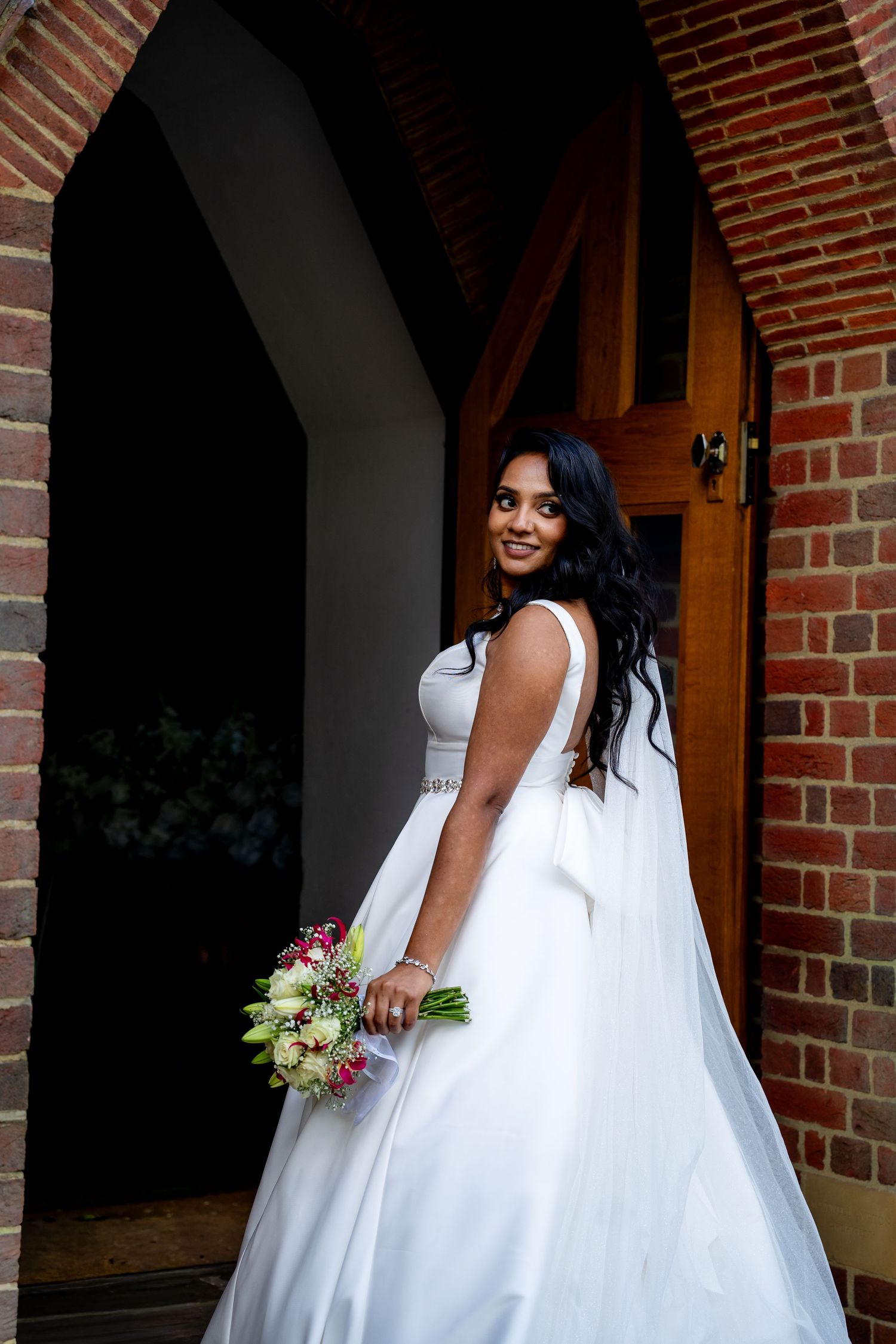 Beautiful bride in white wedding gown holding floral bouquet standing in brick church doorway.
