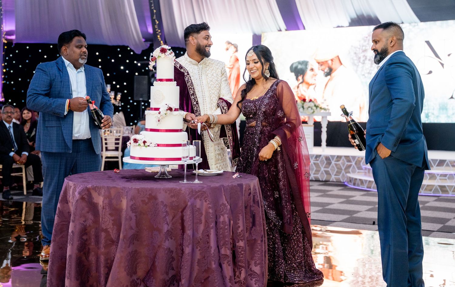 Bride and groom cutting a tall white wedding cake surrounded by guests at Indian reception.