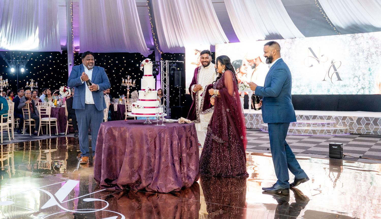 South Asian wedding reception with couple cutting tiered cake, guests in formal attire on decorated dance floor.
