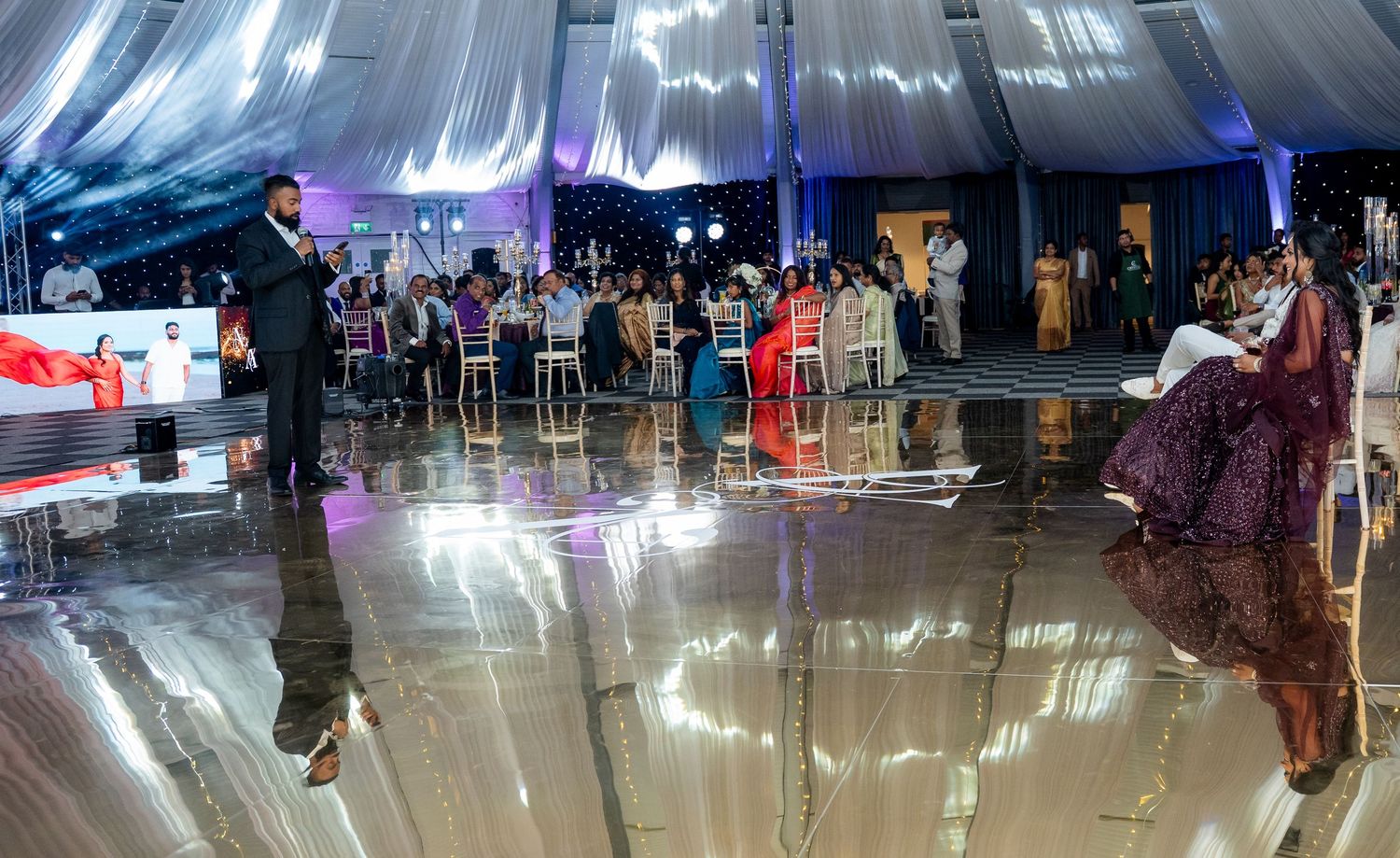 Elegant wedding reception in a decorated tent with guests, shiny dance floor, and a man standing near the stage.