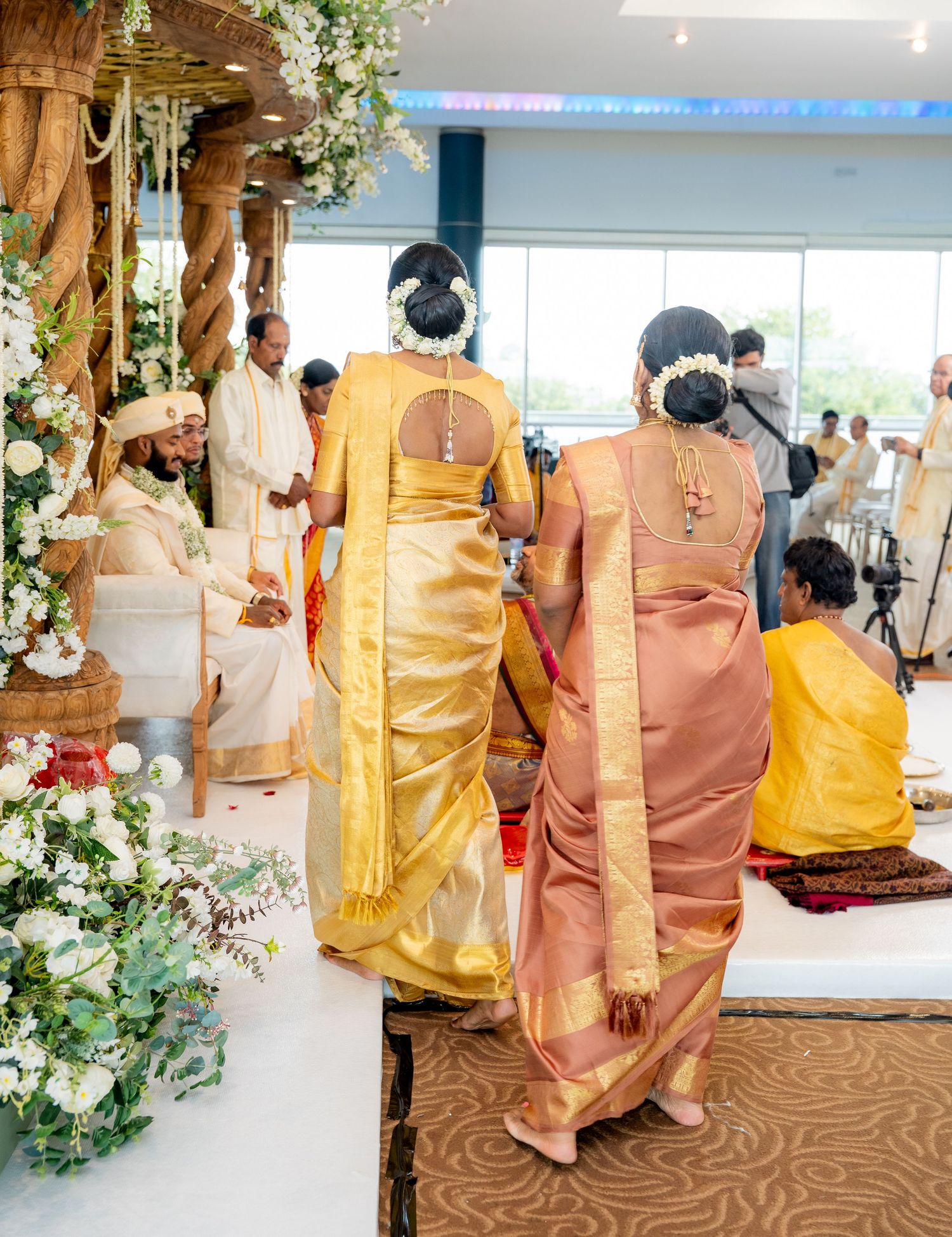 Two women in gold silk sarees walk toward a decorated mandap during a traditional South Indian wedding ceremony.