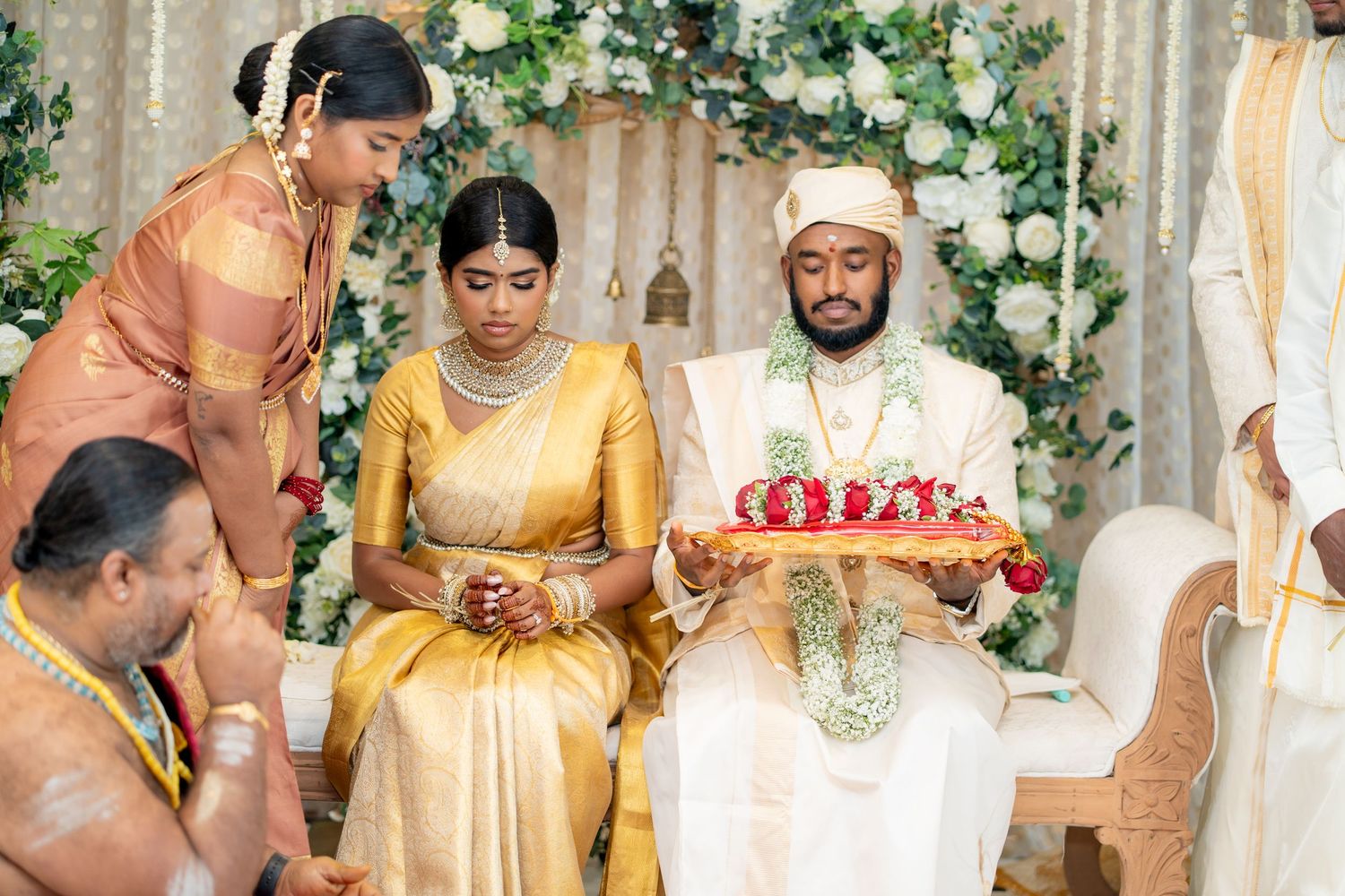 Traditional South Indian wedding ceremony with bride and groom in gold attire, exchanging rituals with flower decorations.
