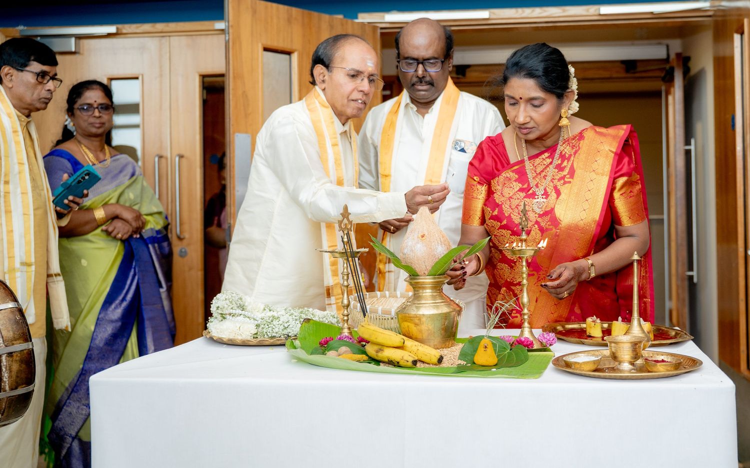 Family members in traditional Indian clothing perform a puja ceremony with brass vessels and banana offerings.