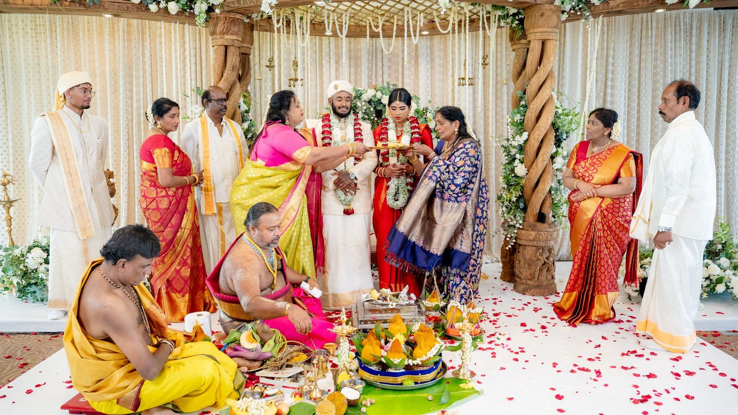 Traditional South Indian wedding ceremony with bride in red saree and groom in white exchanging garlands on decorated mandap.