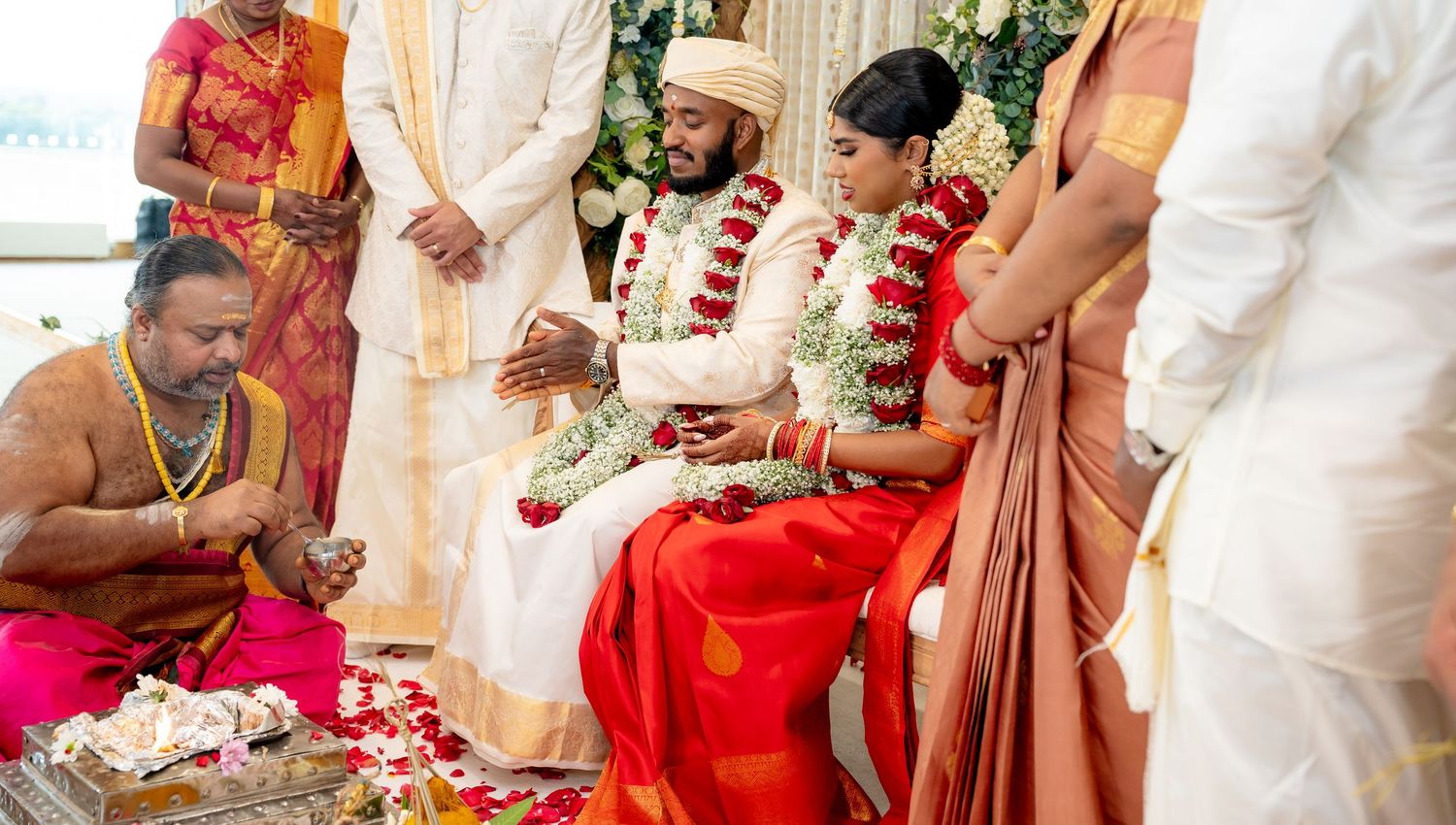 Priest performs traditional Hindu wedding rituals for seated couple adorned with flower garlands on mandap.
