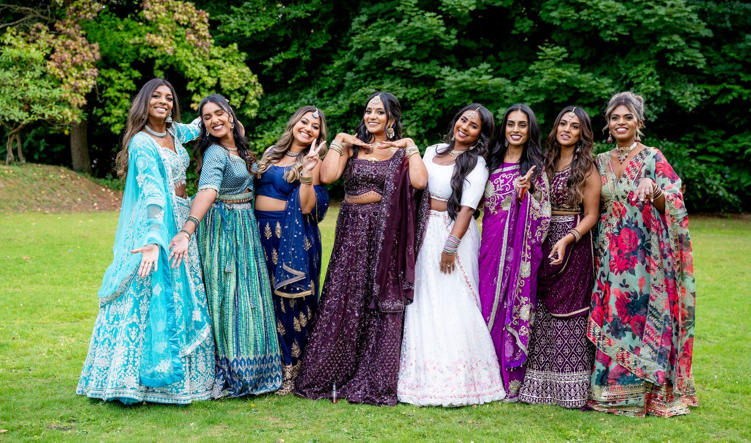 Group of women in vibrant Indian attire posing outdoors on green grass with lush trees in background.