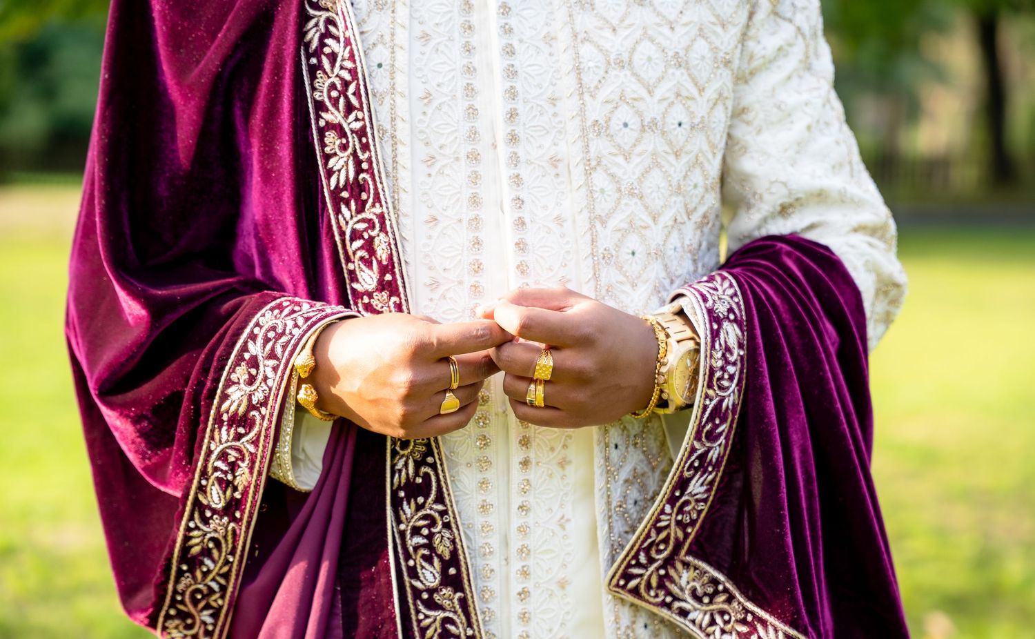 Close-up of groom in white sherwani with burgundy velvet embroidered dupatta and gold bangles at Indian wedding.