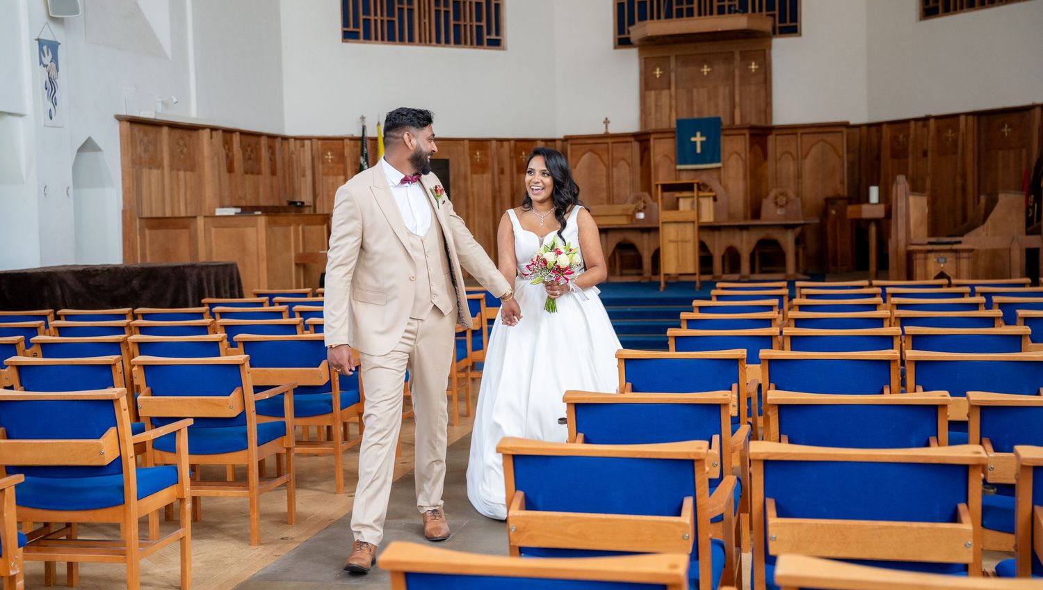 Newlywed couple walking hand in hand between blue pews inside a wooden church.