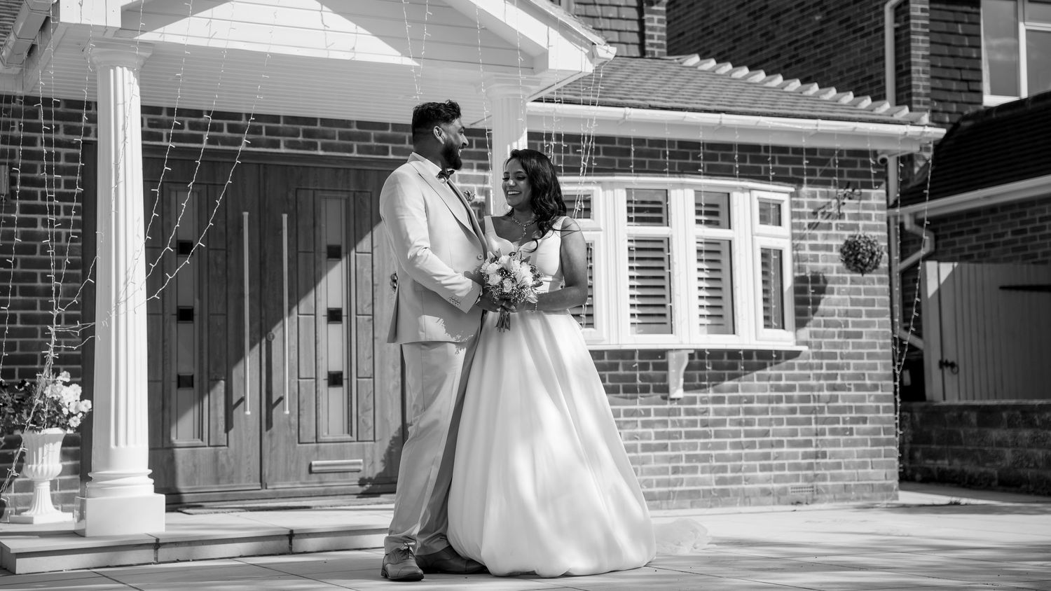 Black and white photo of a bride and groom posing together outside a brick building on their wedding day.