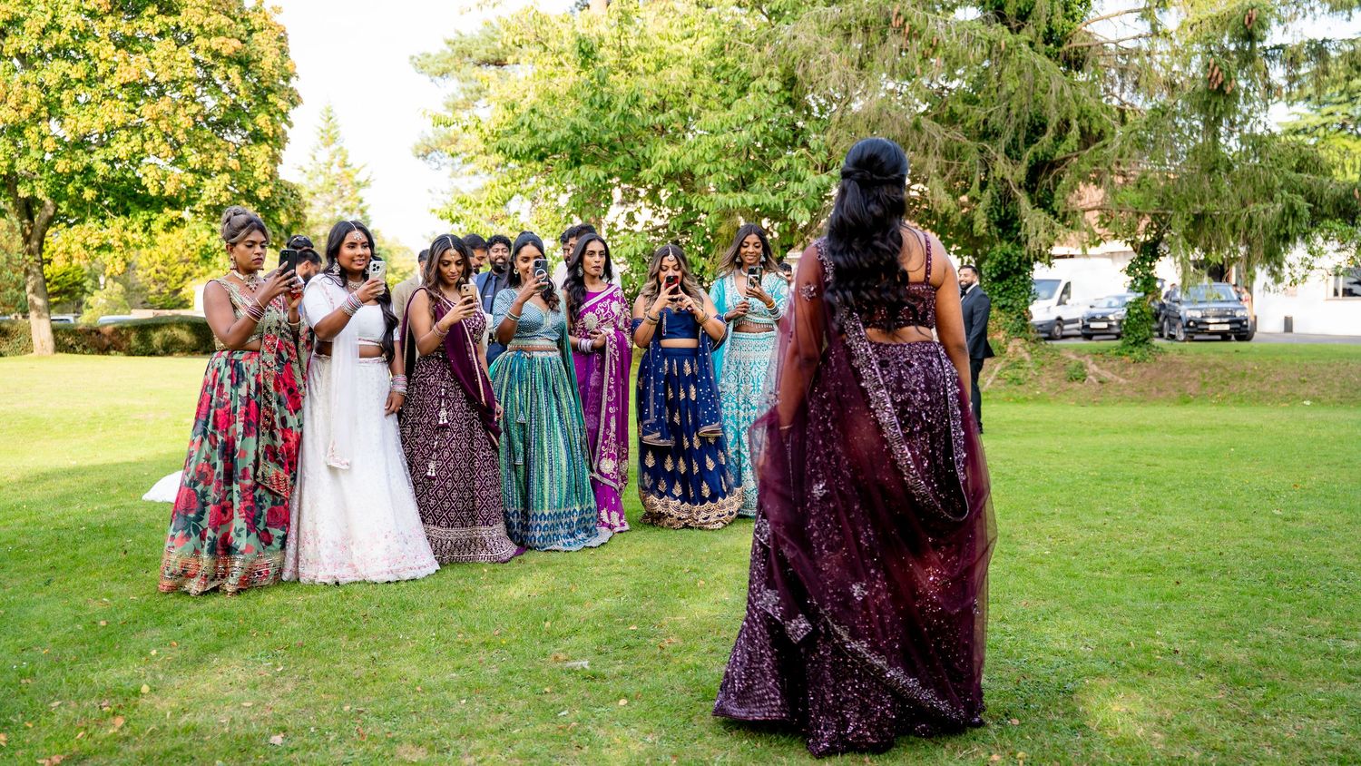Indian bride in maroon lehenga faces bridesmaids in colorful sarees on green lawn at outdoor wedding celebration.