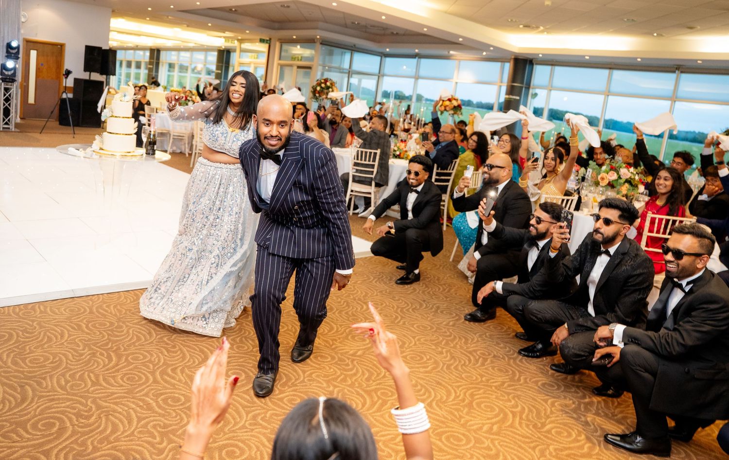 Groom in pinstripe suit dances with bride in silver lehenga before seated wedding reception guests.