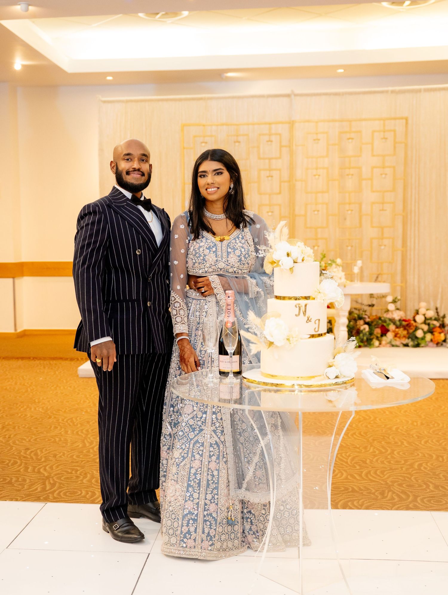 Couple poses in wedding attire beside tiered white and gold cake at reception venue.