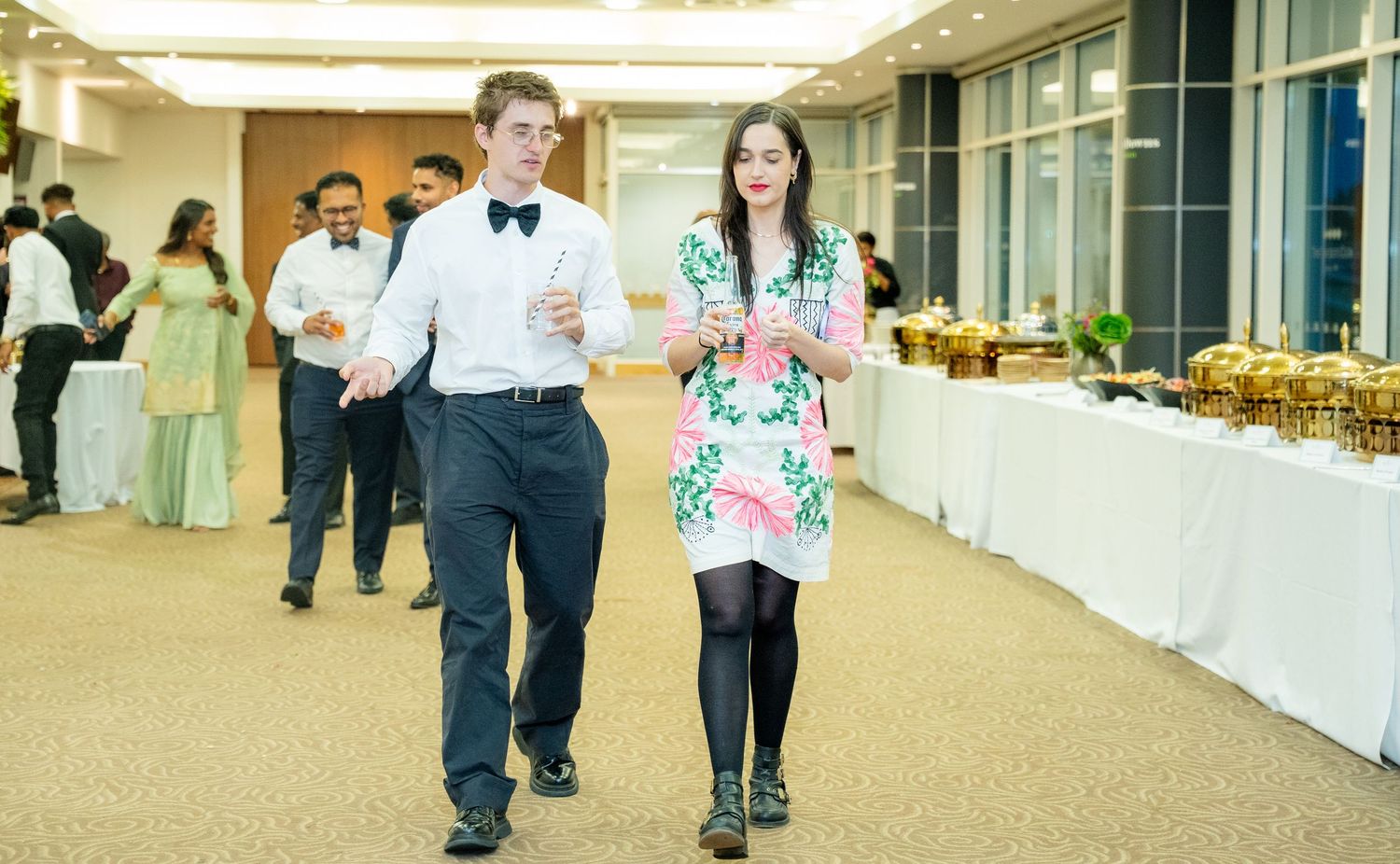 Young couple in formal wear walking through a grand banquet hall with buffet display.