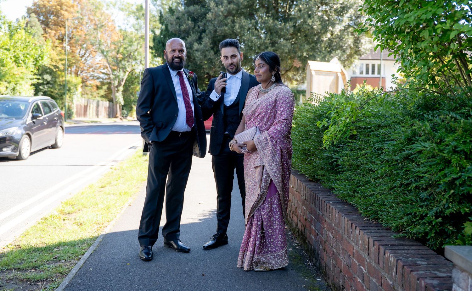 Wedding guests in formal attire including woman in pink saree posing on street before ceremony.