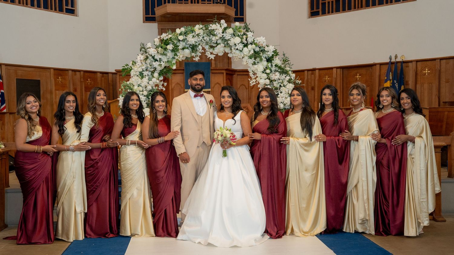 Bride and groom pose with bridesmaids in burgundy and gold gowns under floral arch in elegant church wedding ceremony.