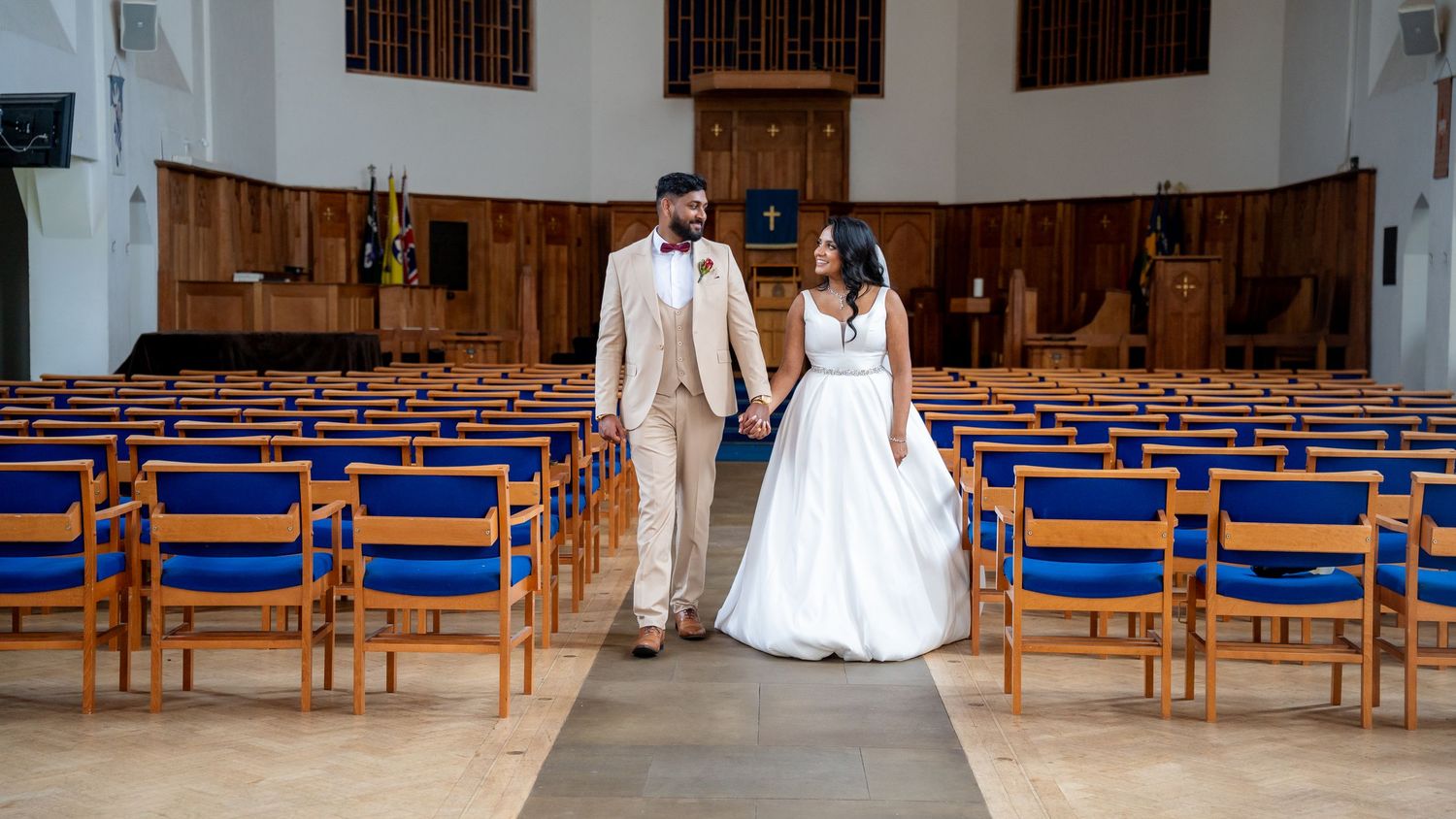Bride and groom holding hands walking down the aisle of a church with blue chairs during their wedding ceremony.