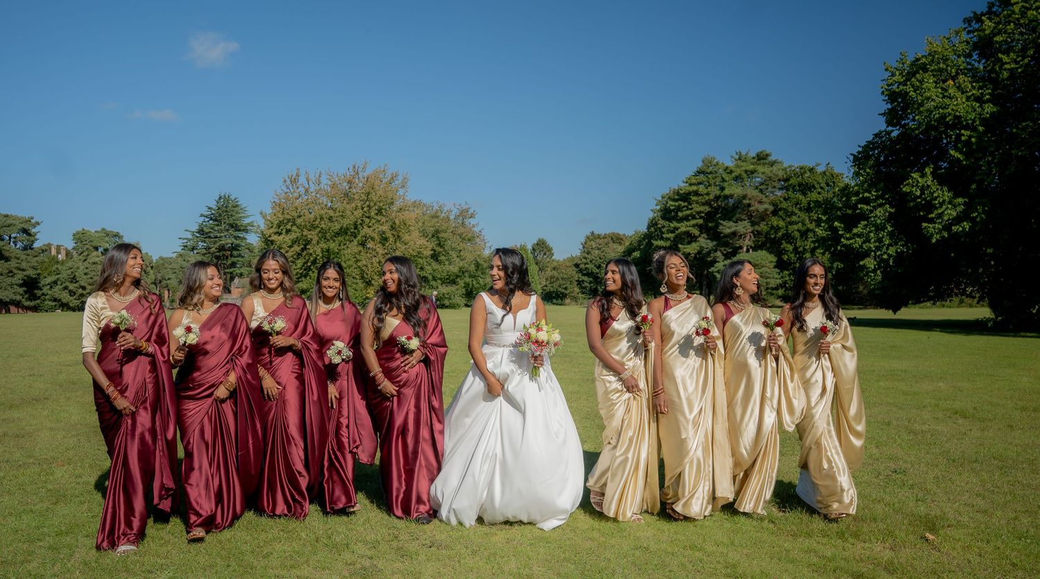 Bride in white gown walks with bridesmaids in burgundy dresses and gold sarees outdoors.
