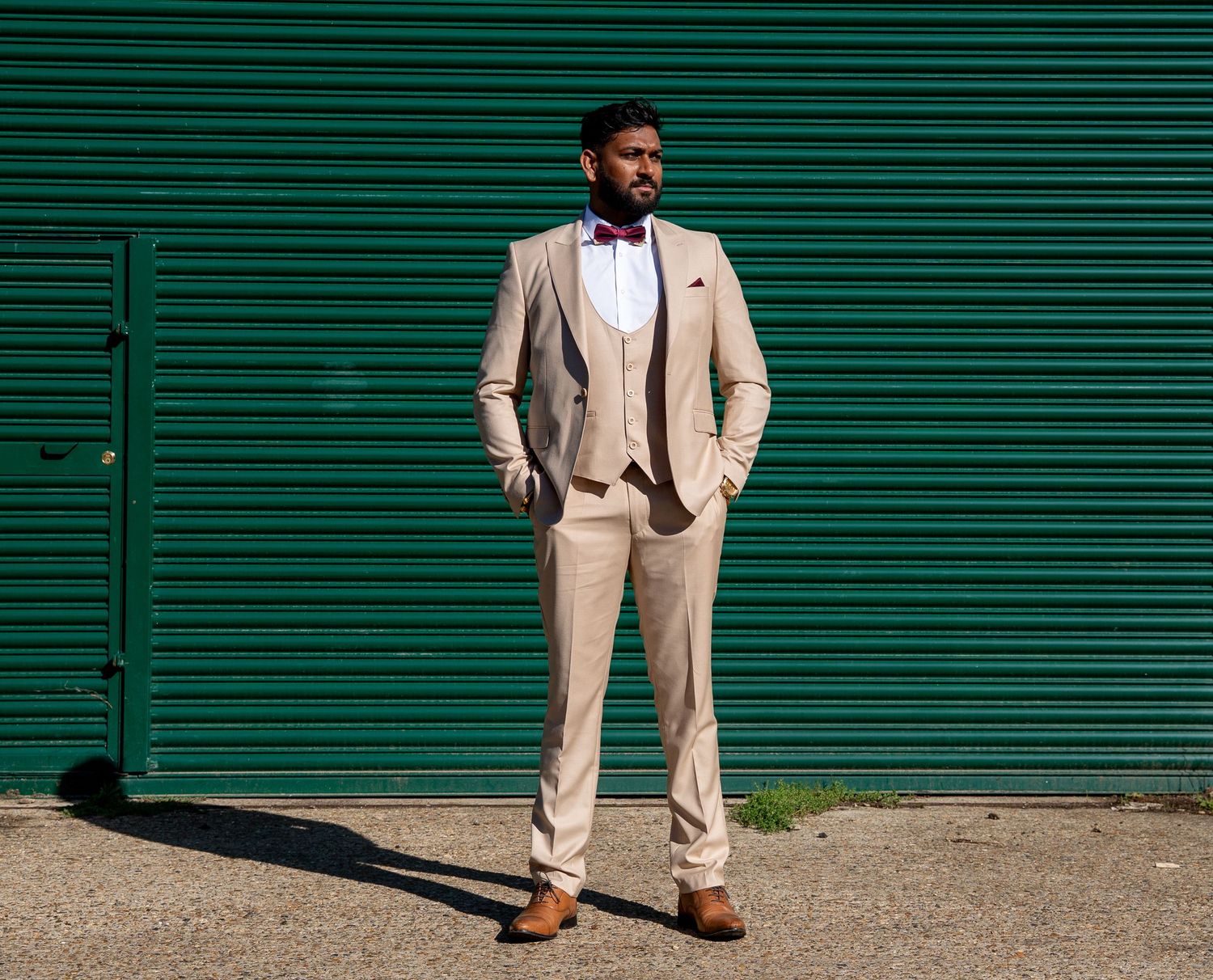 Groom in beige three-piece suit with burgundy bow tie poses confidently against a green shutter.